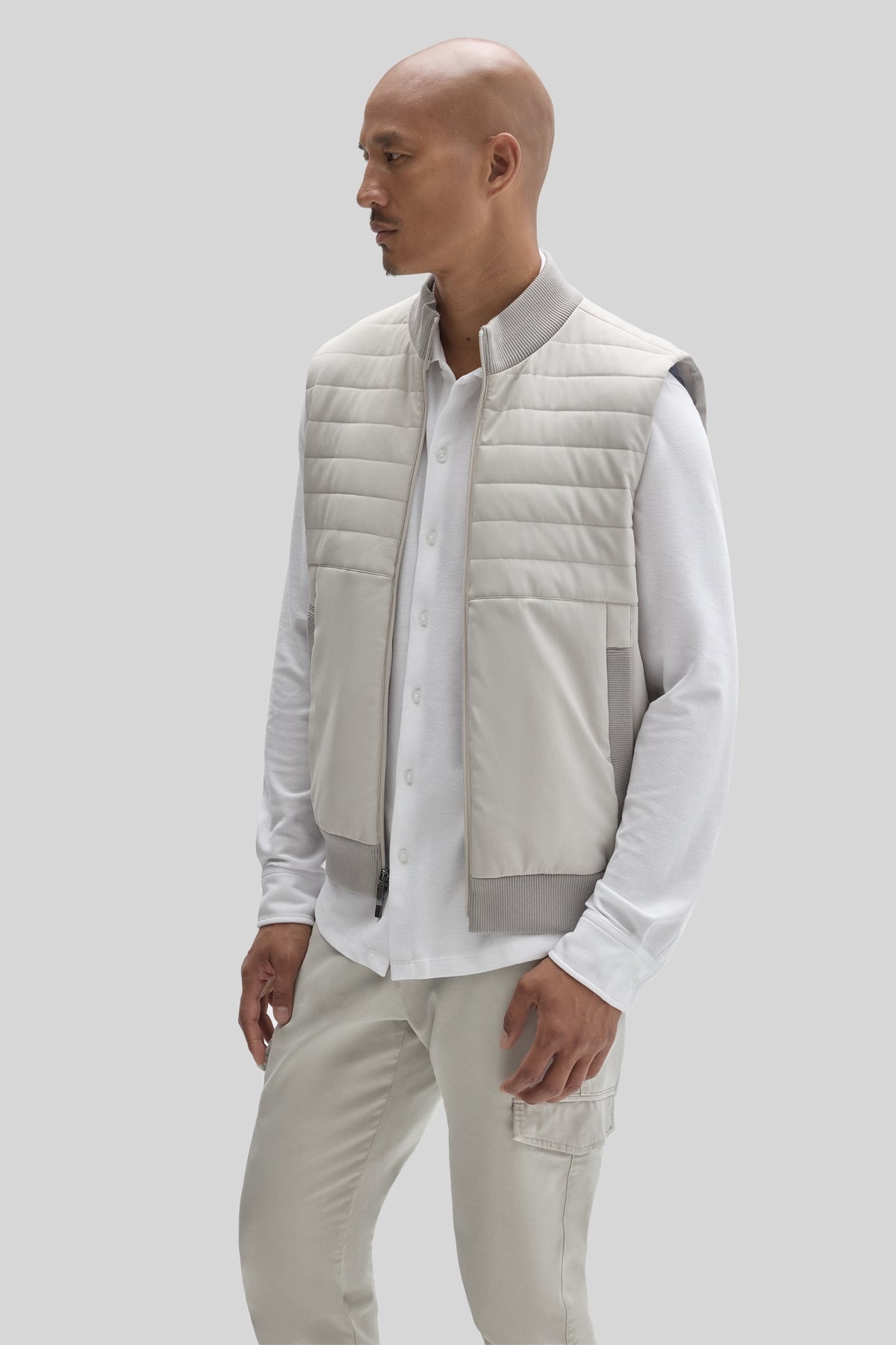 Beige quilted vest with horizontal stitching, ribbed collar and hem, worn over white long-sleeve button-up shirt.