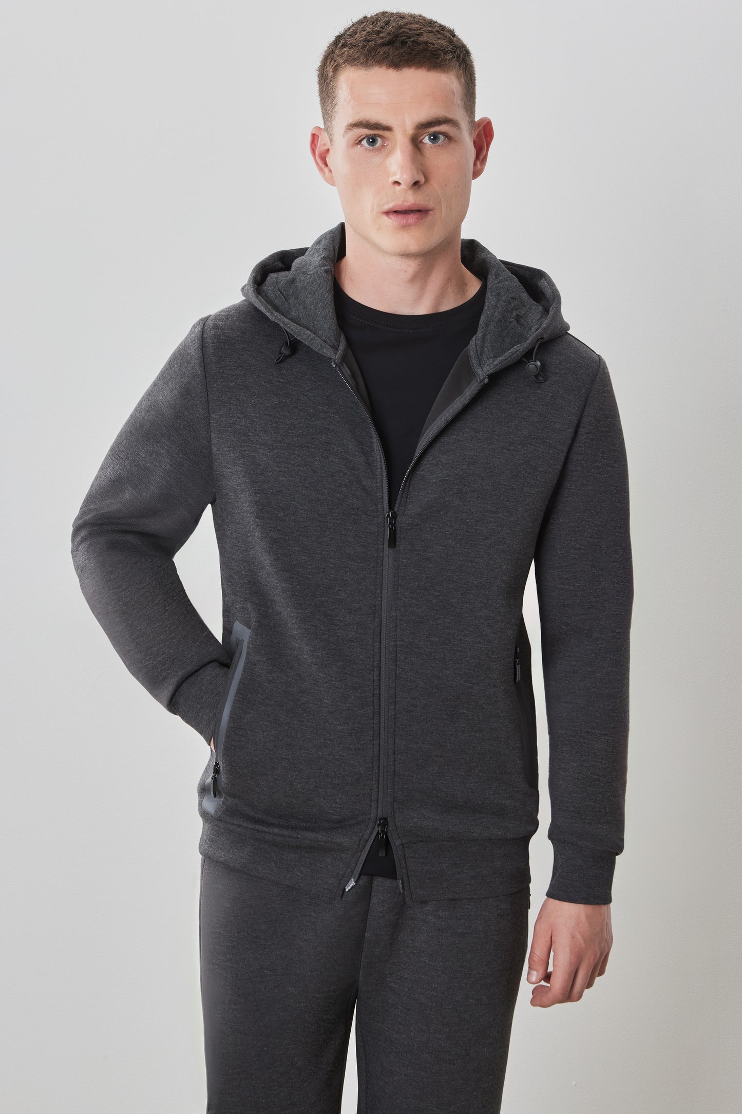 A male model wearing a long-sleeve full zip hoodie in charcoal color, with the hood on and a front zipper pocket.