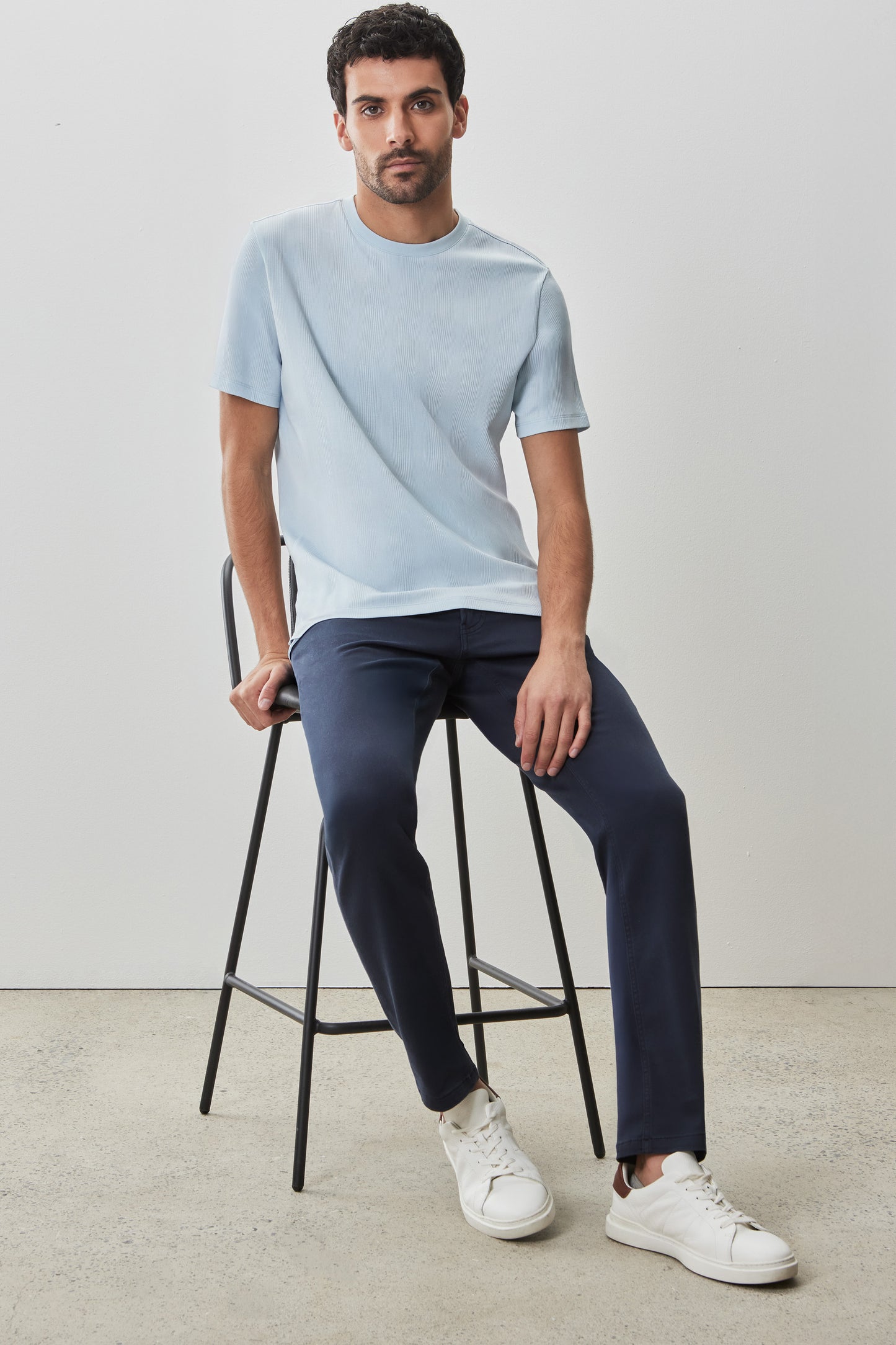 Man wearing light blue textured short-sleeve crew neck tee with dark navy pants and white sneakers, seated on a black stool.
