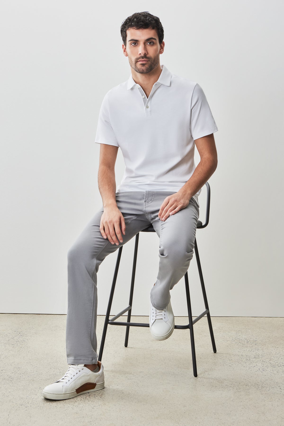 Man wearing white short-sleeve polo shirt with collar and gray pants, seated on a stool.