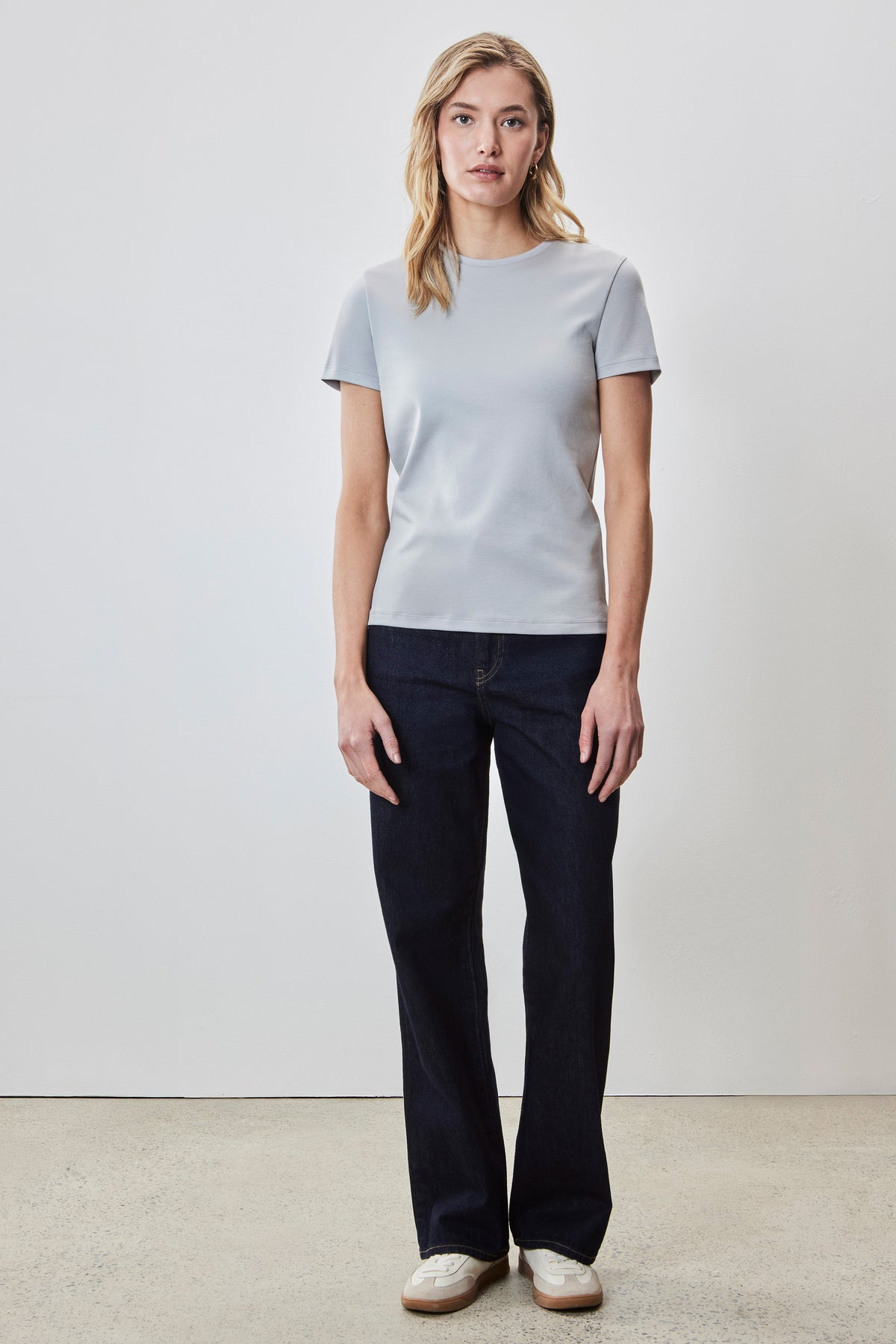 Woman wearing light gray short-sleeve crew neck tee with dark indigo jeans and white sneakers.