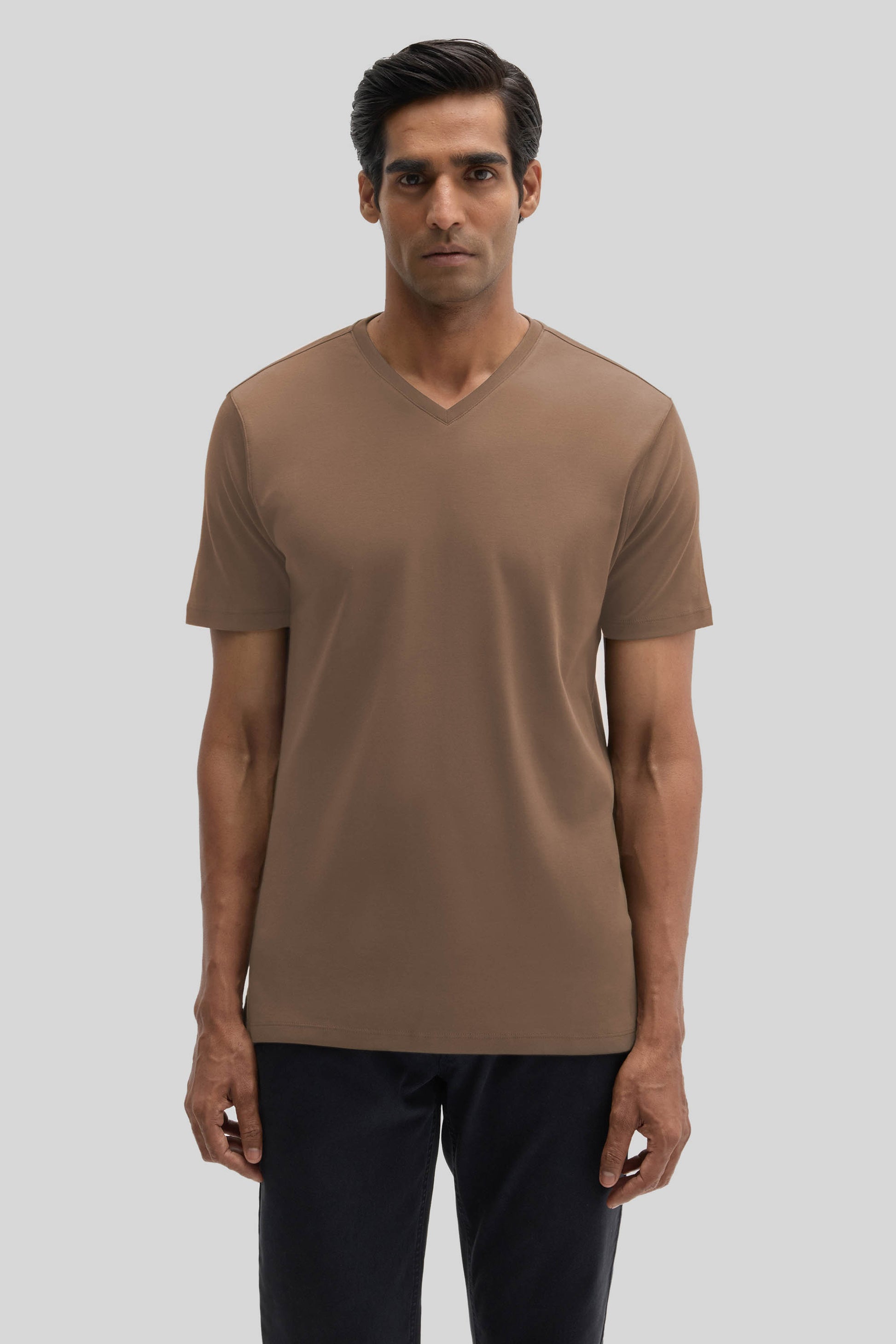 Man wearing a taupe V-neck t-shirt with short sleeves and dark pants against a neutral background.