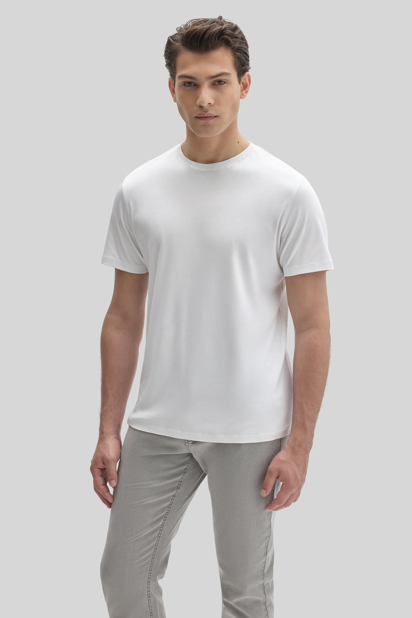 Man wearing white crew neck short-sleeve t-shirt with gray pants against neutral background.
