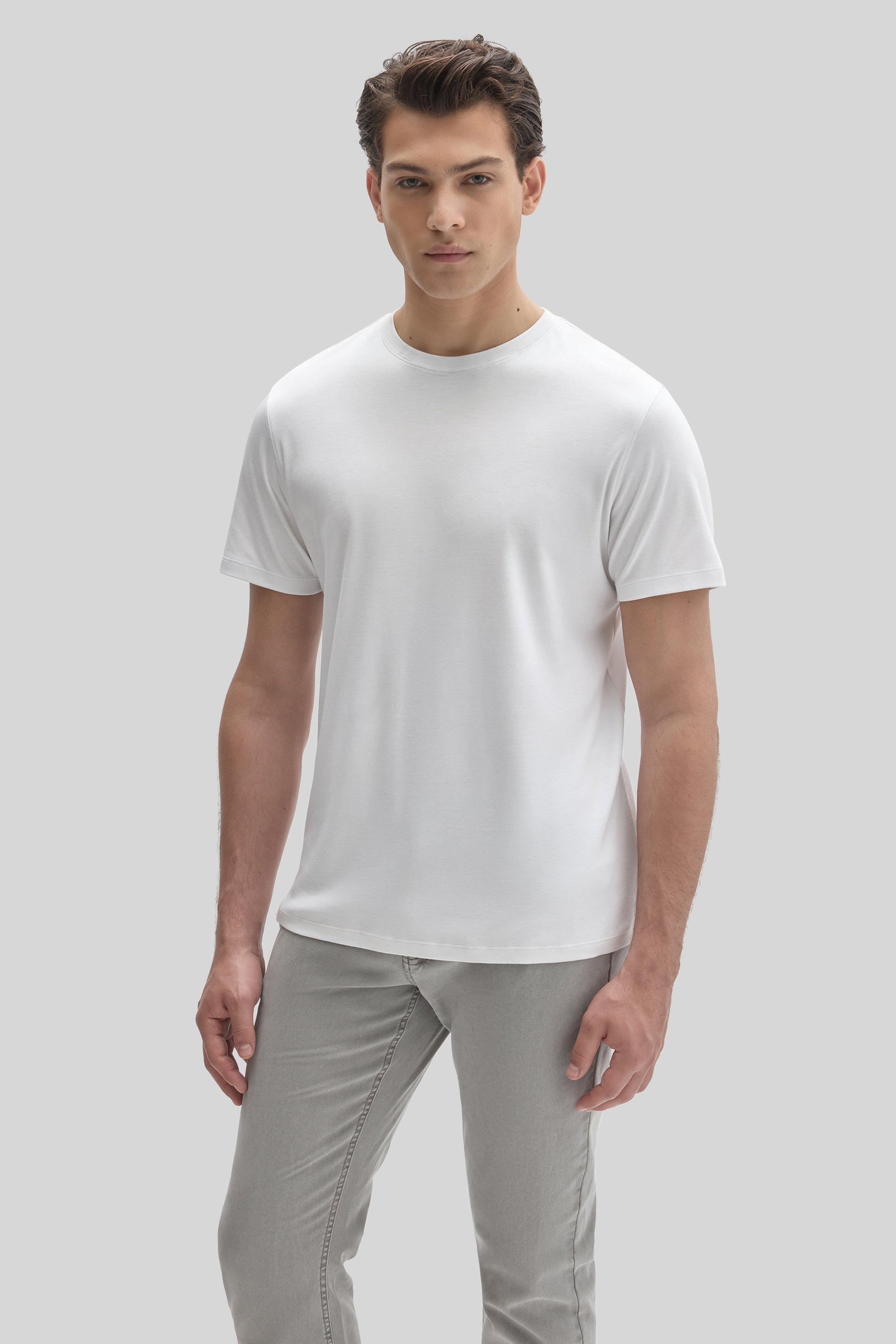 Man wearing white crew neck short-sleeve t-shirt with gray pants against neutral background.