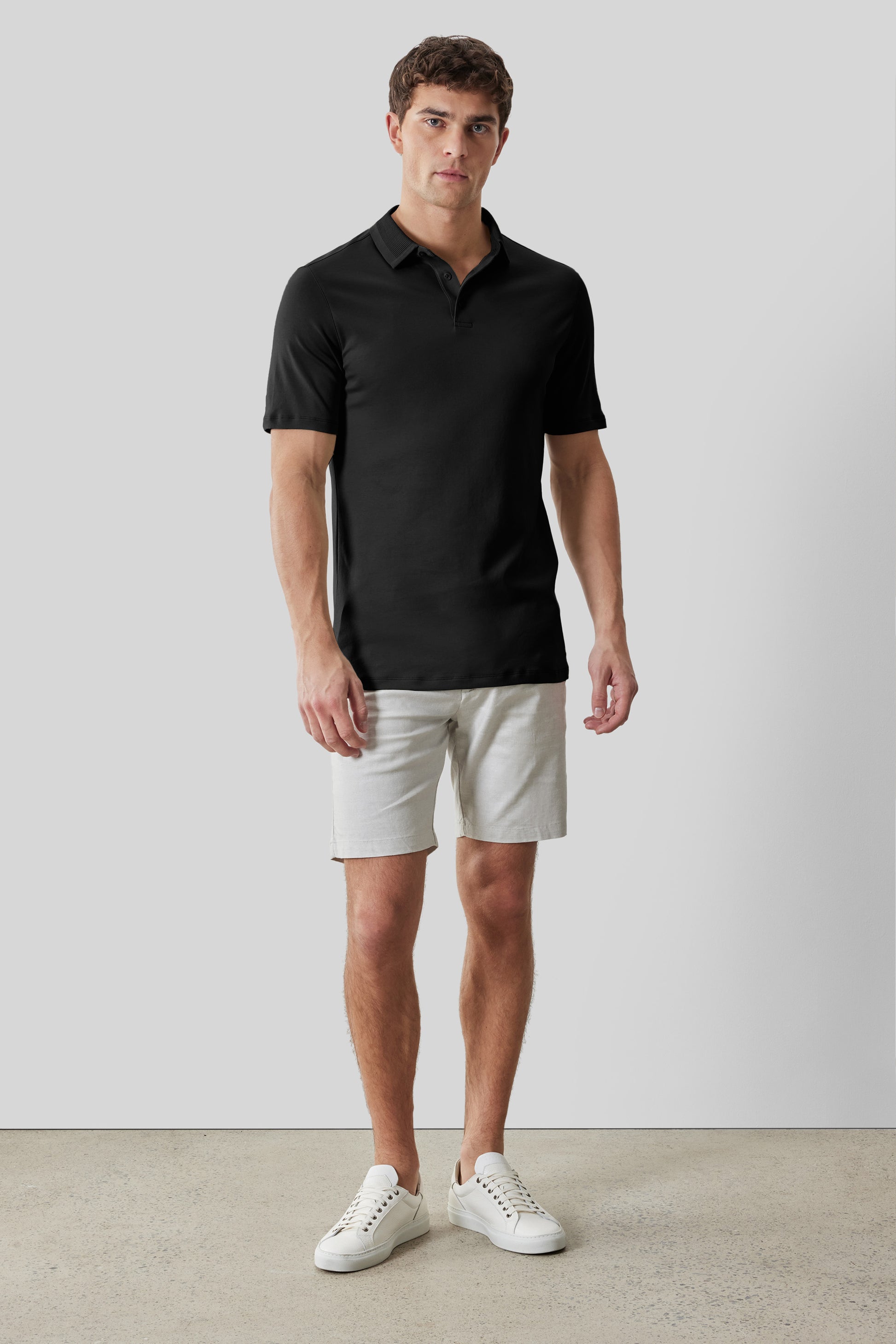Man wearing black short-sleeve polo shirt with textured collar and beige shorts paired with white sneakers.