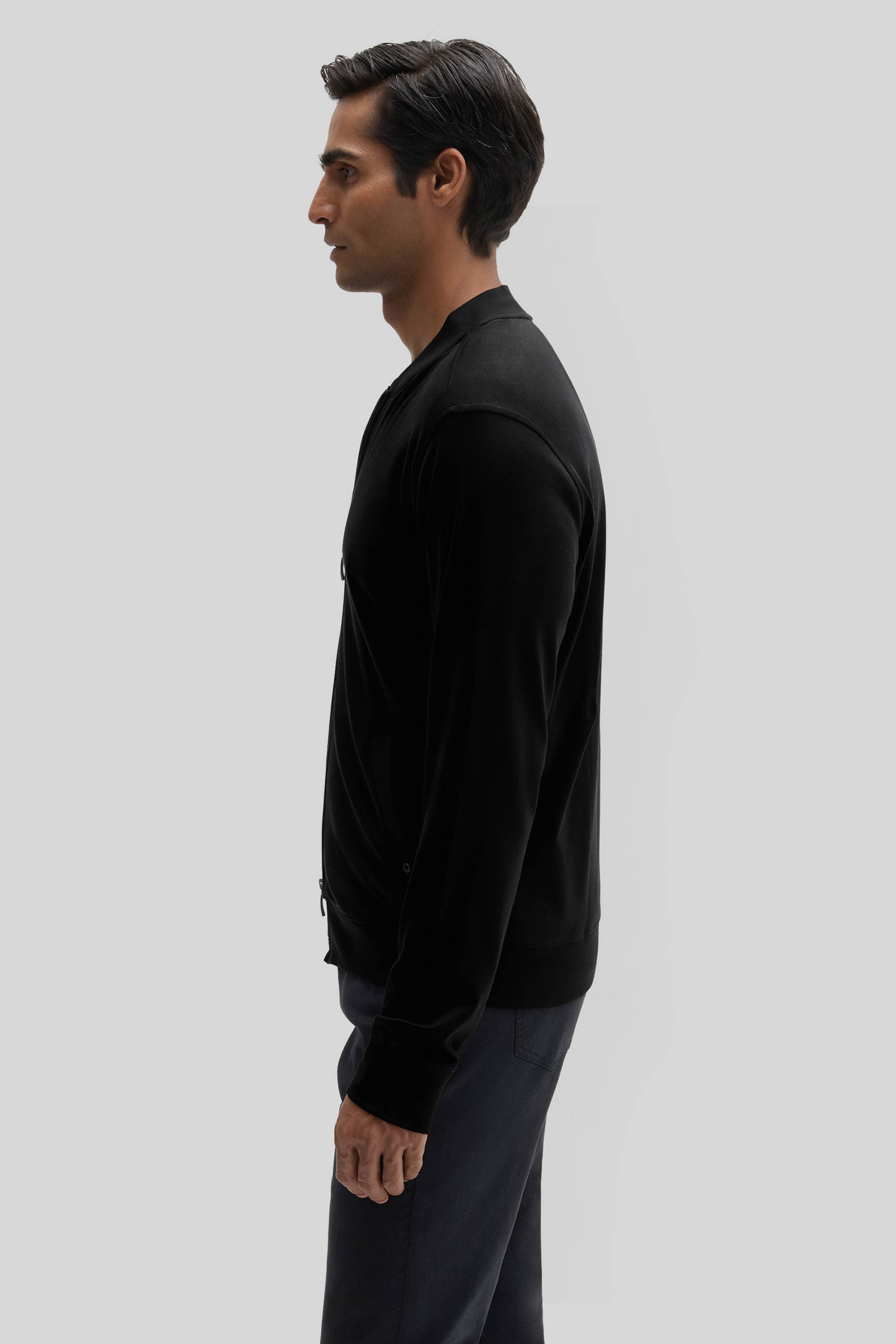Man wearing a black baseball jacket with ribbed cuffs and dark pants, shown in side profile.