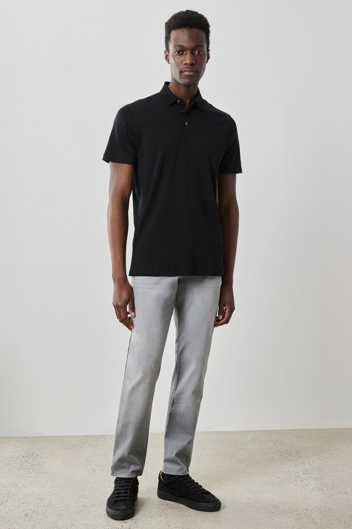 Man wearing black short-sleeve polo shirt with collar and buttons, paired with light gray denim jeans and black sneakers.
