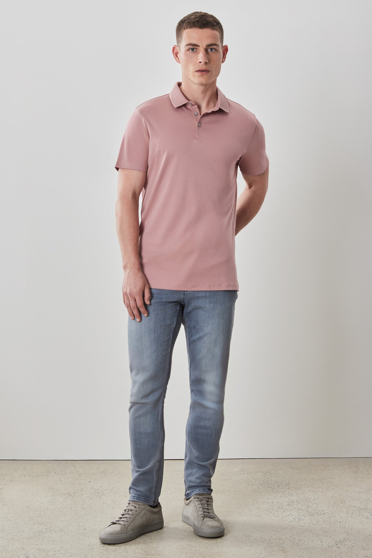 Man wearing a mauve pink polo shirt with blue jeans and gray sneakers.