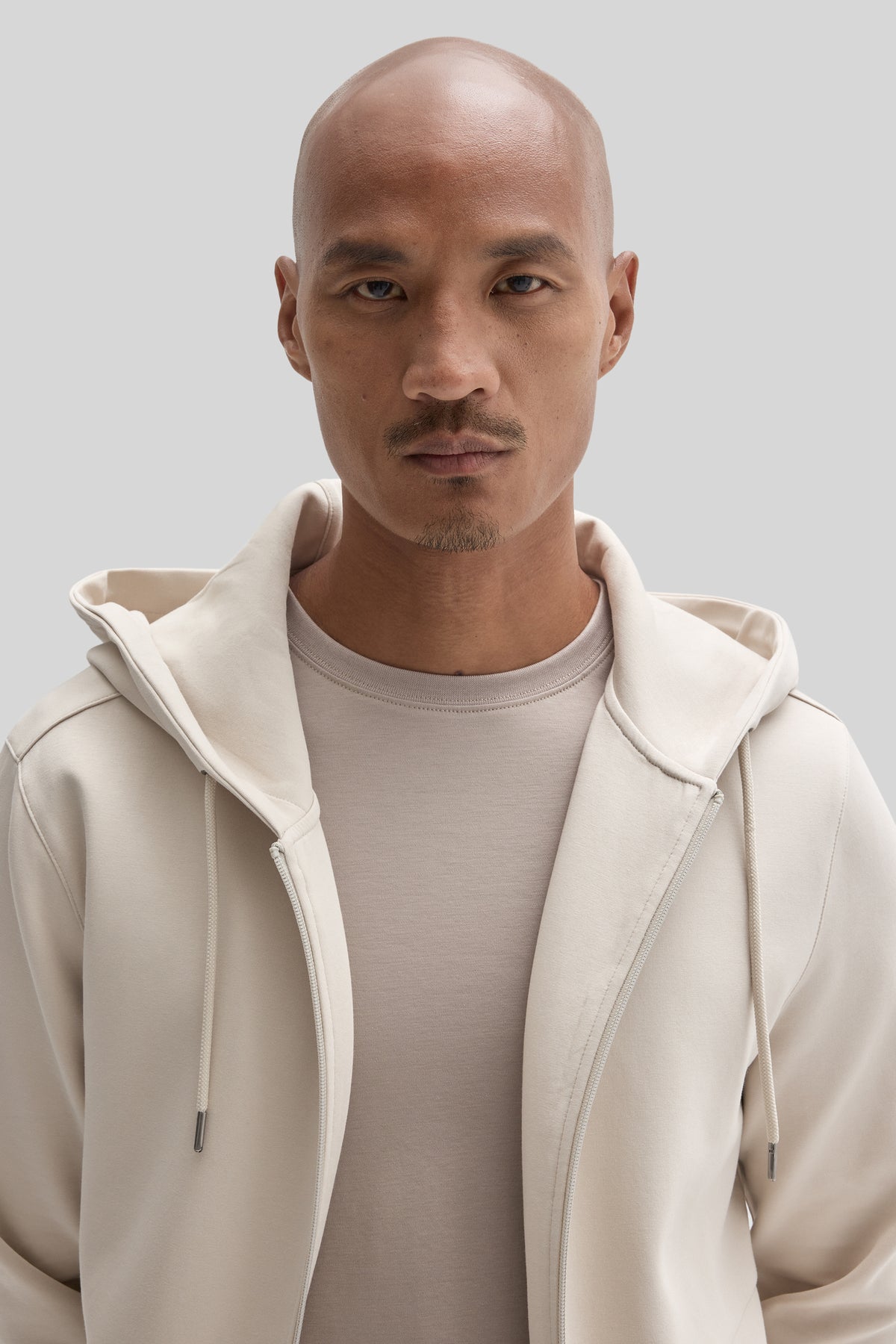Man wearing cream full zip hoodie over gray t-shirt against neutral background.