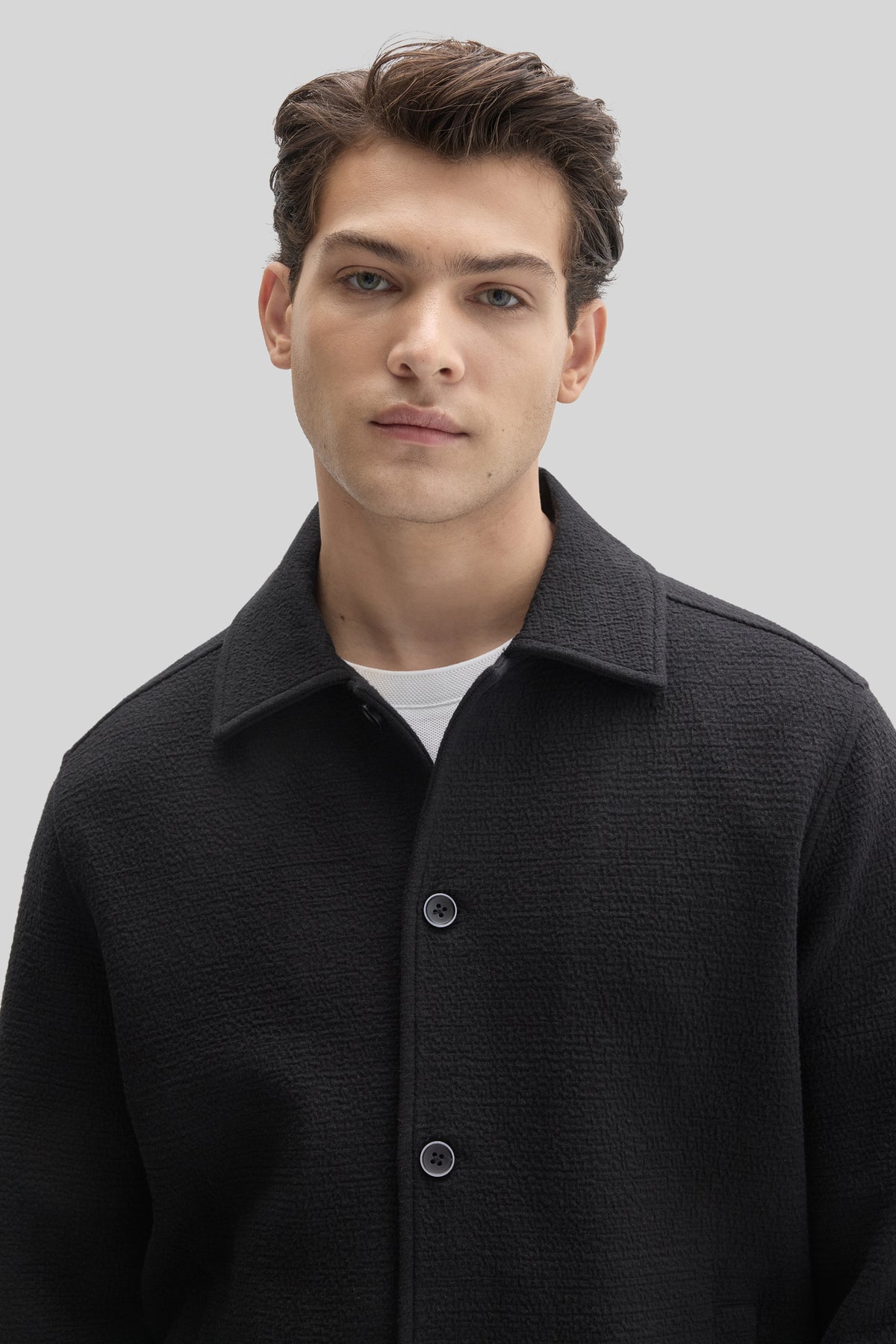 Man wearing black textured buttoned jacket with collar over white shirt.