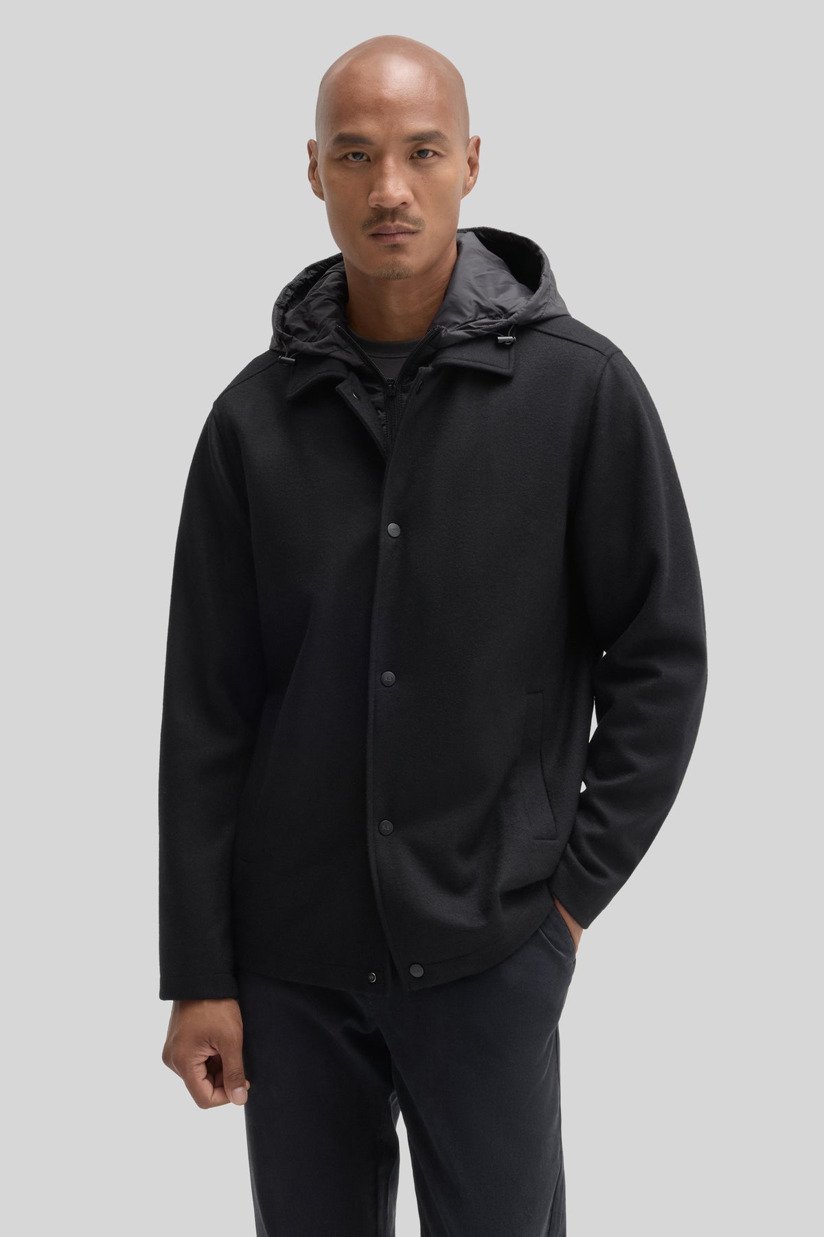 Black button-up car coat with hood and front pockets worn by a man against a neutral background.