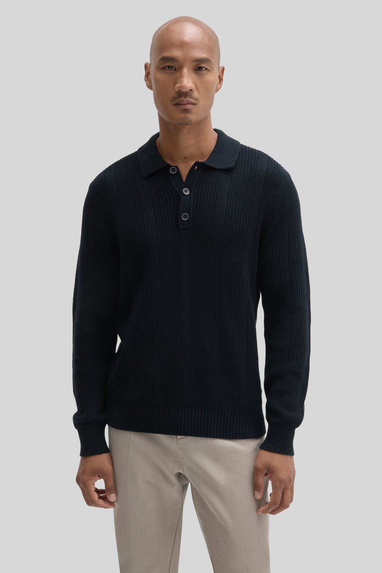 Black ribbed sweater polo with three-button placket and collar, worn with beige trousers.