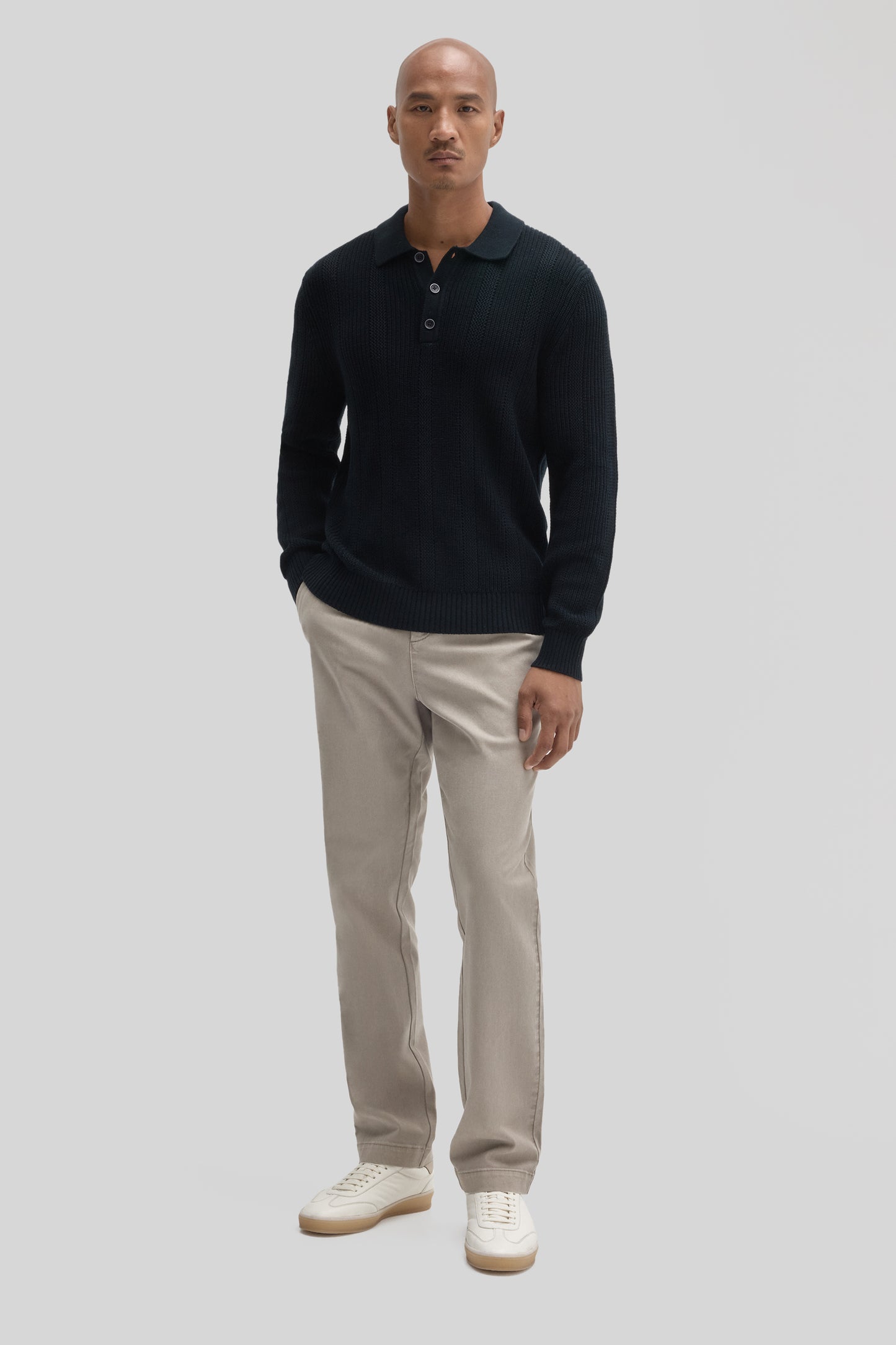 Black ribbed long-sleeve polo sweater with collar and button front, worn with beige pants and white sneakers.