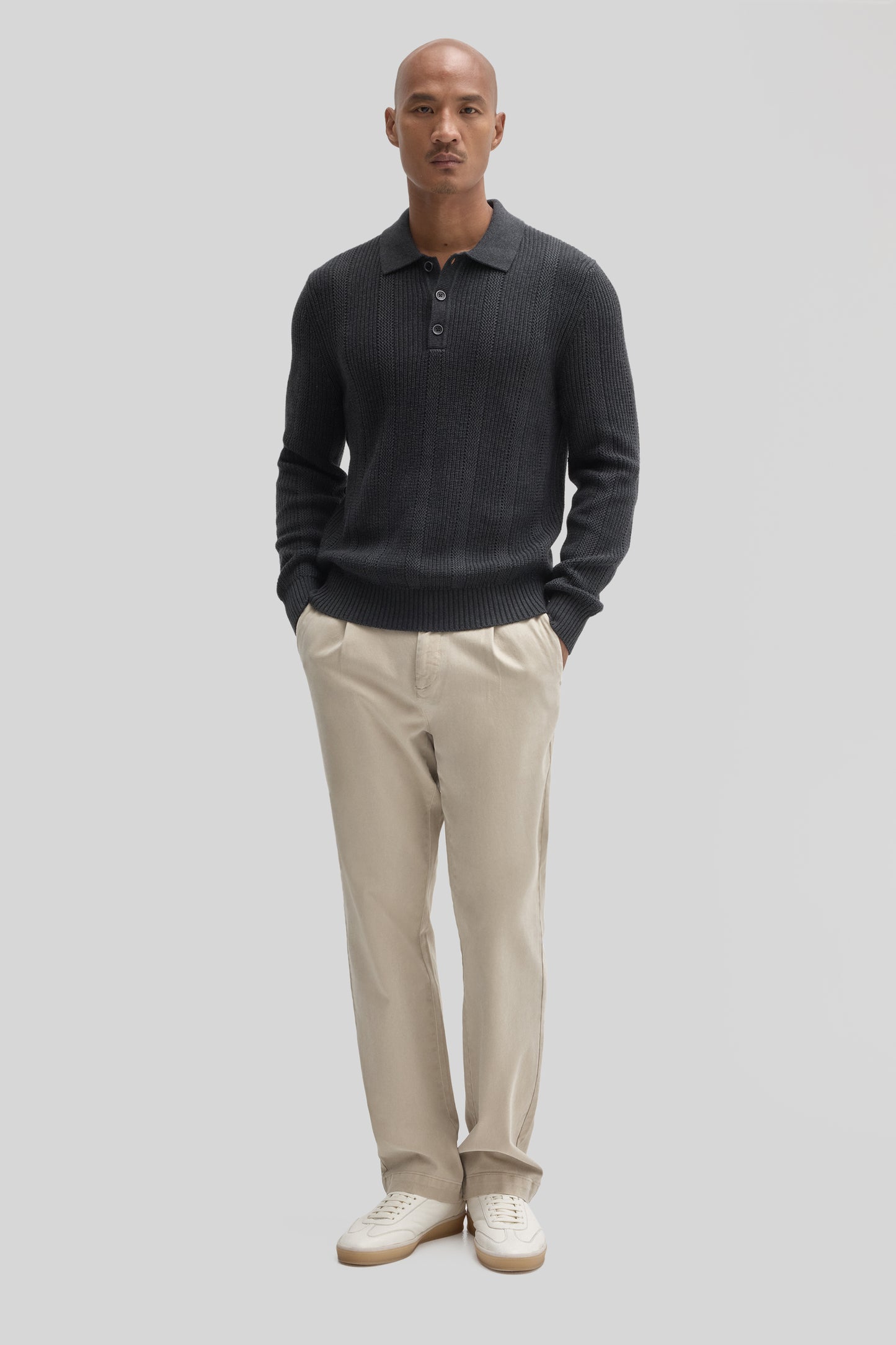 Man wearing dark gray ribbed polo sweater with collar and buttons, paired with beige chinos and white sneakers.