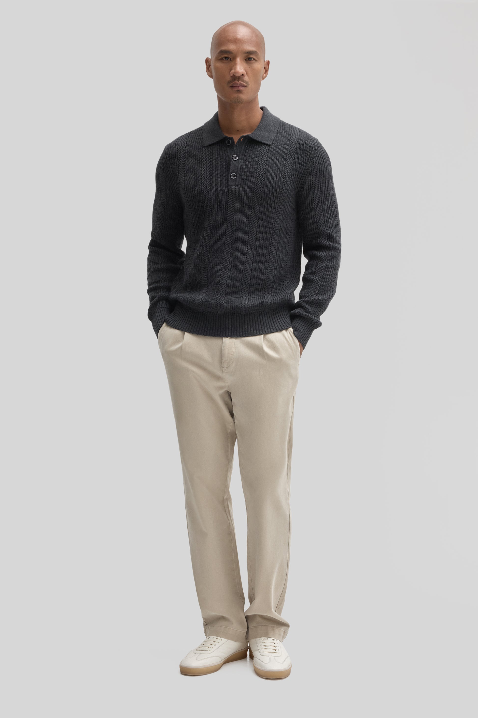 Man wearing dark gray ribbed polo sweater with collar and buttons, paired with beige chinos and white sneakers.