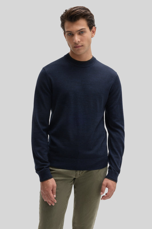 Man wearing a navy crewneck sweater with long sleeves and khaki pants against a neutral background.