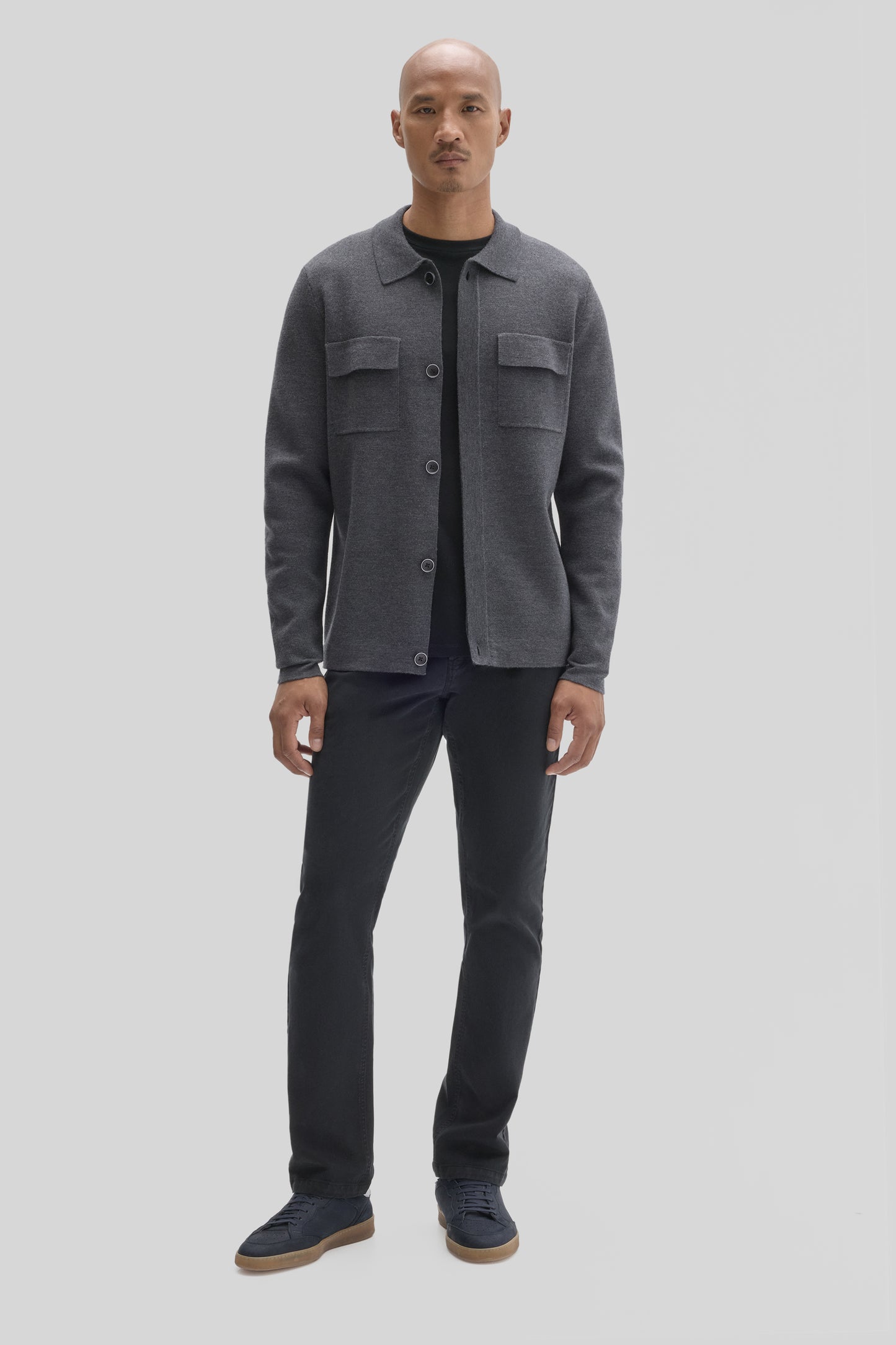 Man wearing charcoal gray buttoned cardigan with chest pockets over black shirt, paired with black jeans and navy sneakers.