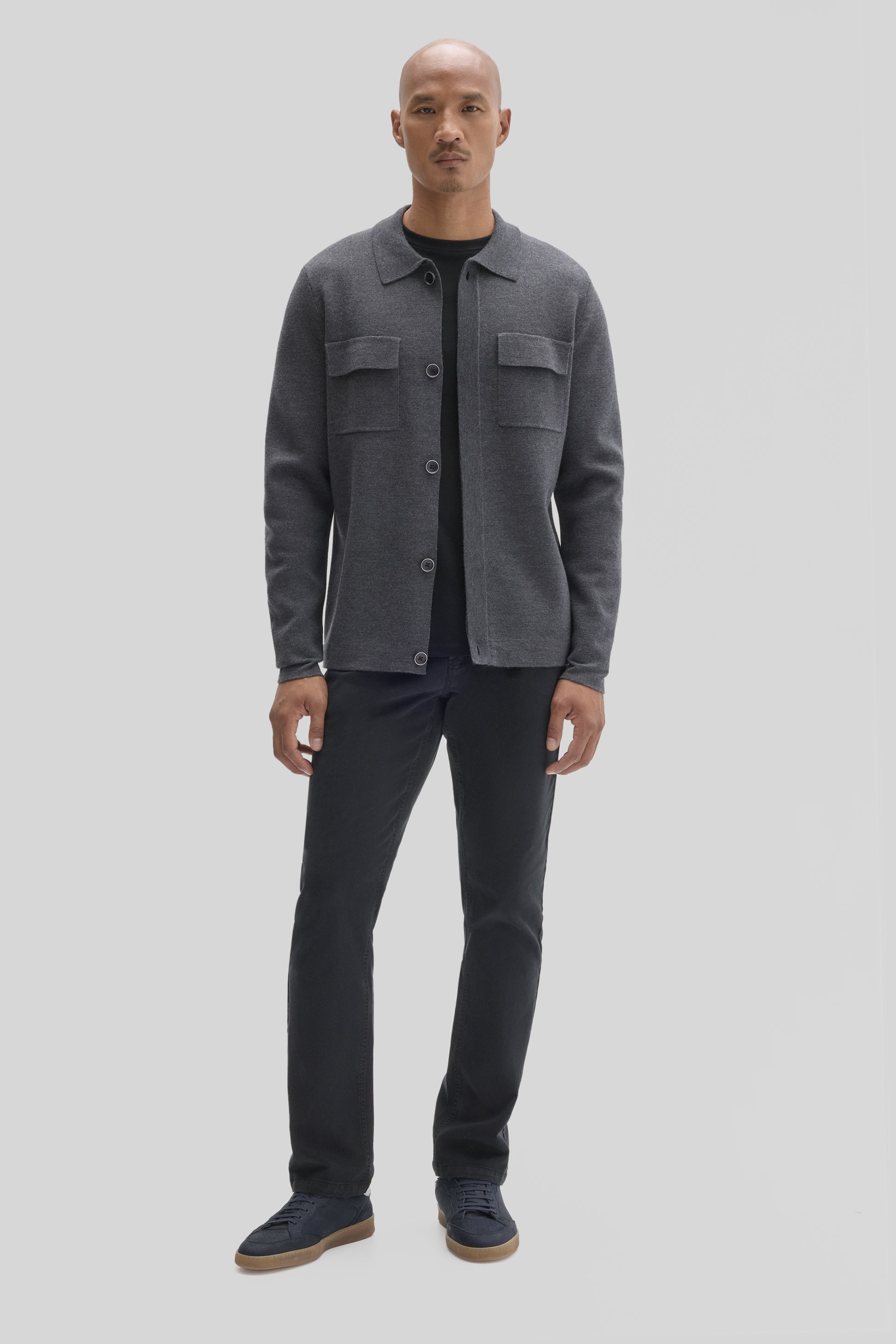 Man wearing charcoal gray buttoned cardigan with chest pockets over black shirt, paired with black jeans and navy sneakers.