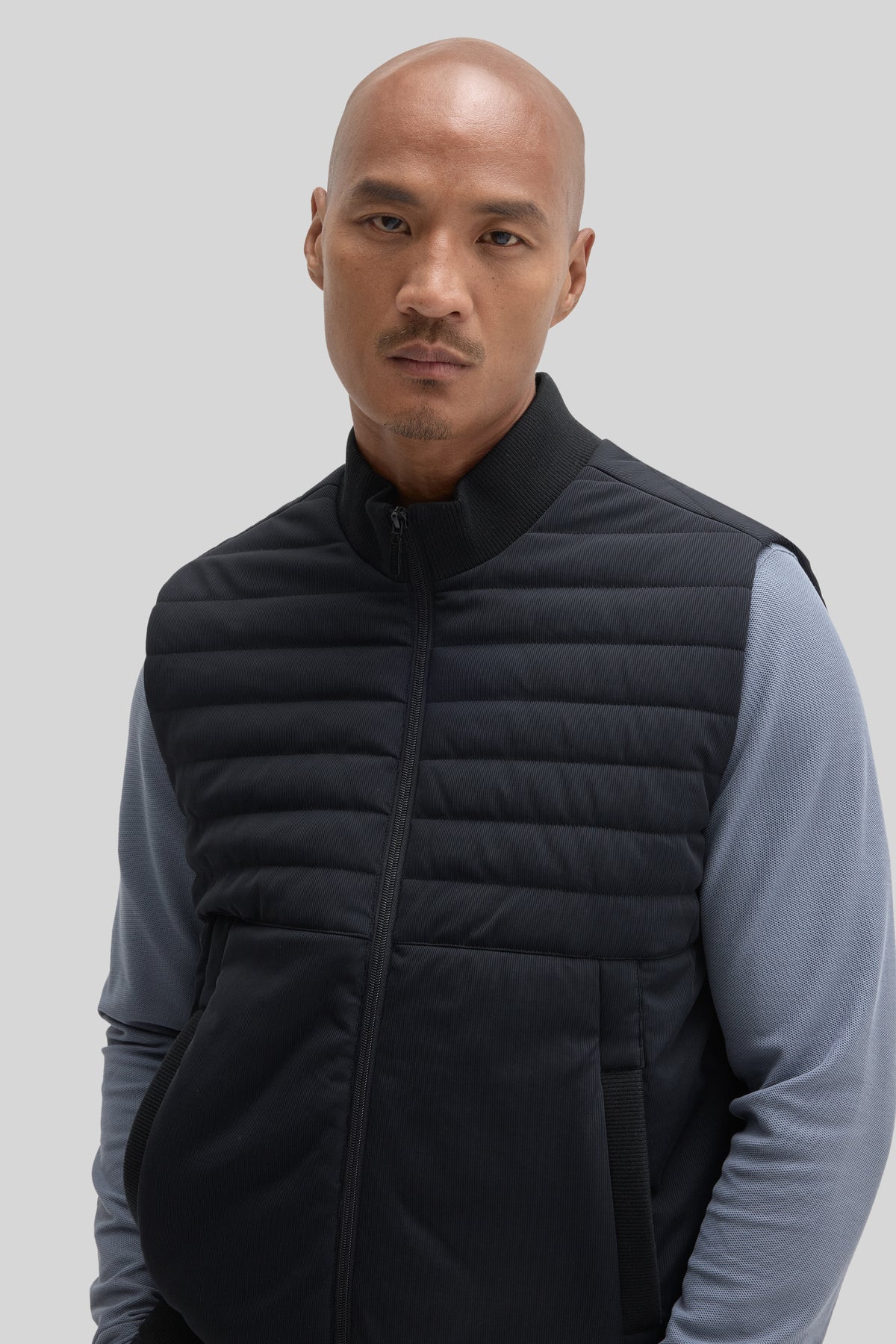 Black quilted vest with horizontal stitching, ribbed collar and armholes, worn over gray long-sleeve shirt.