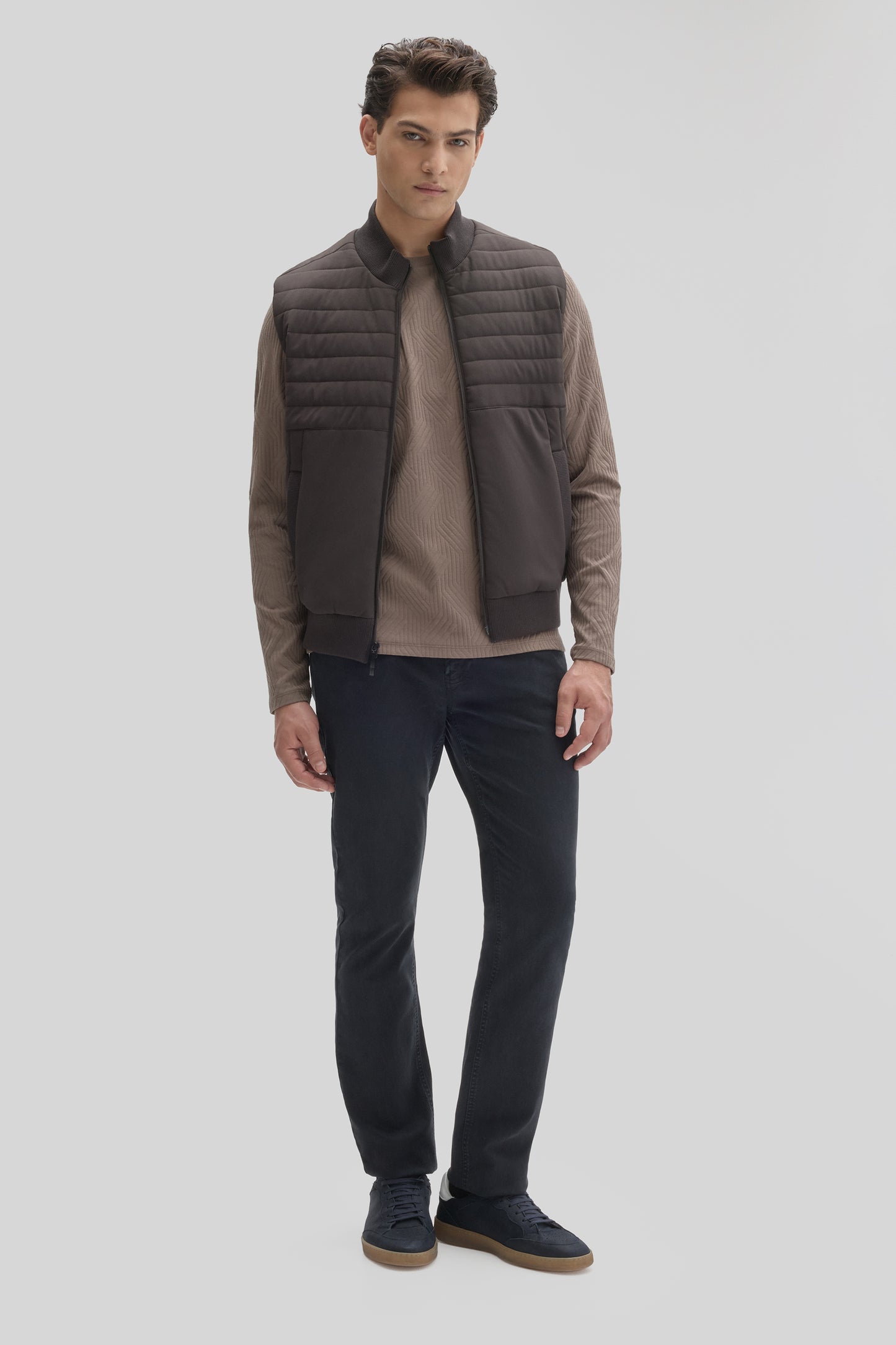 Man wearing a dark gray quilted vest with horizontal stitching over a beige ribbed long-sleeve shirt and navy pants.