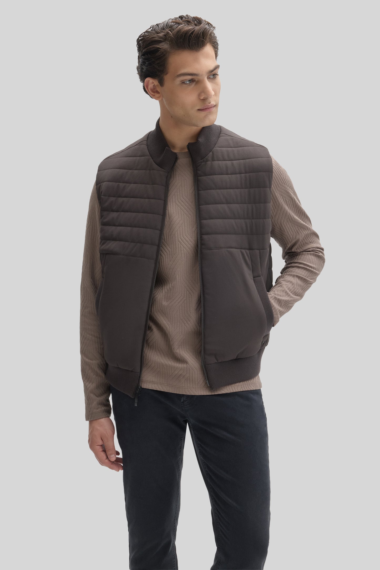 Man wearing dark gray quilted vest with high collar over beige ribbed long-sleeve shirt and navy pants.