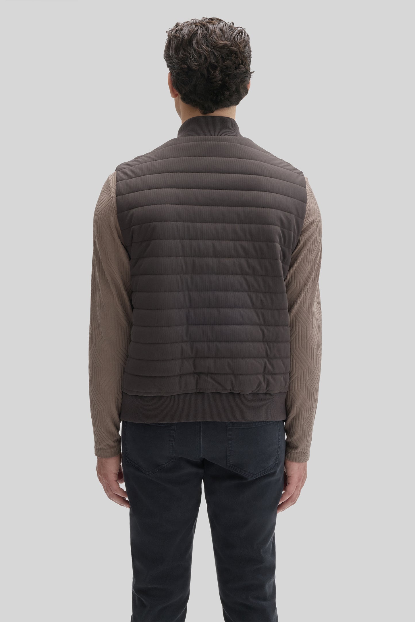Dark gray quilted vest with horizontal stitching, ribbed collar and hem, worn over a tan long-sleeve shirt and dark jeans.