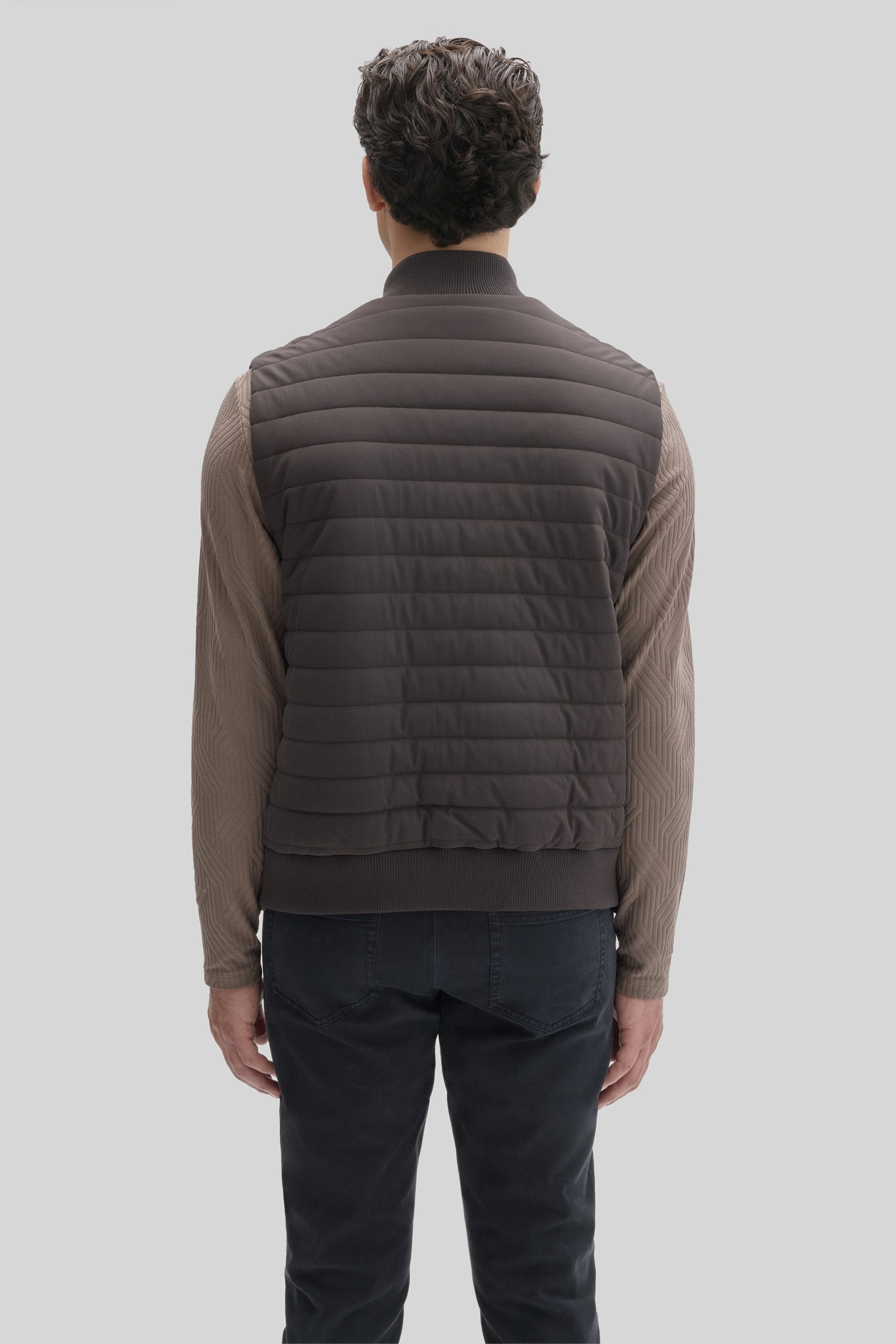 Dark gray quilted vest with horizontal stitching, ribbed collar and hem, worn over a tan long-sleeve shirt and dark jeans.