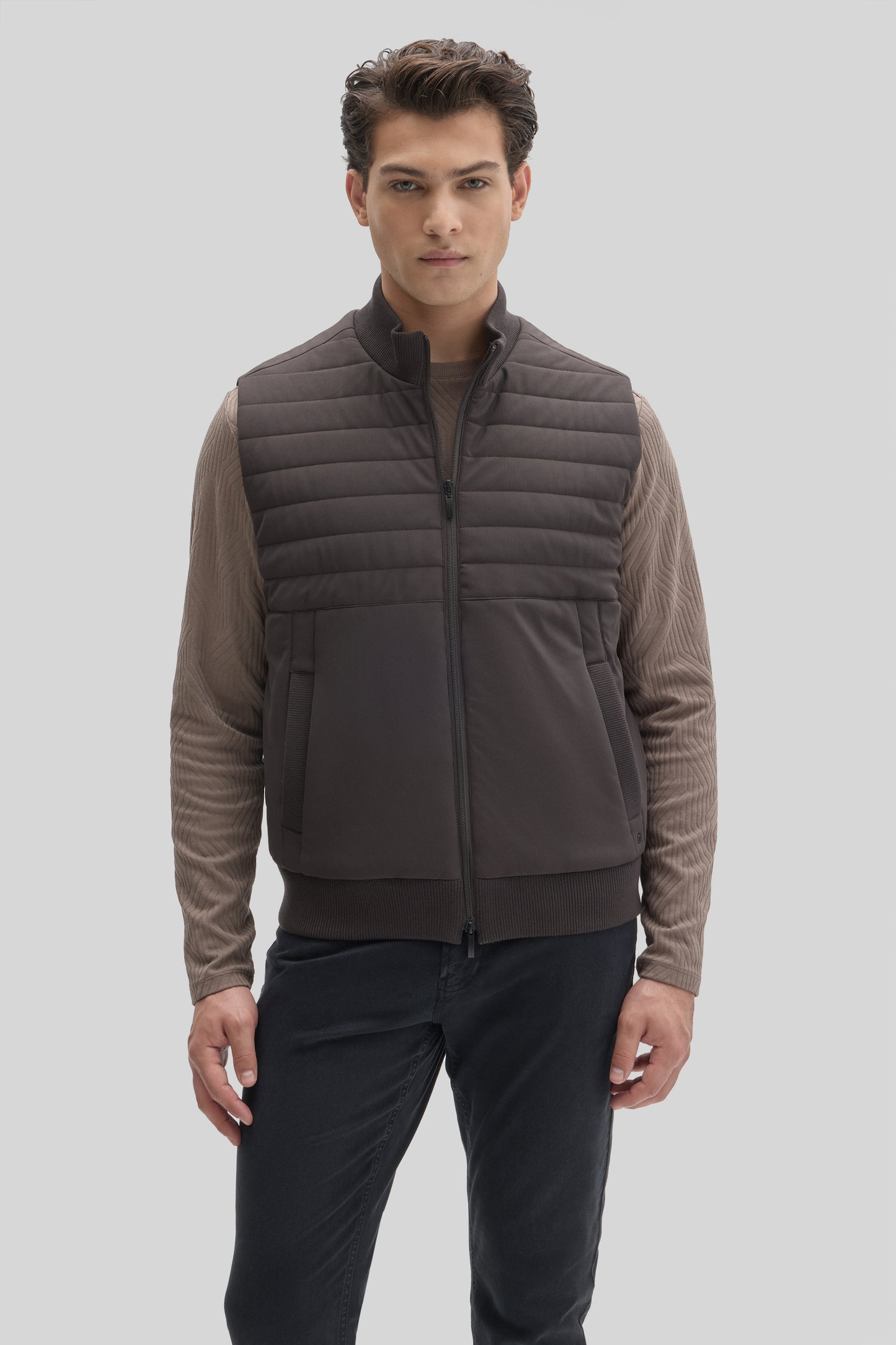 Man wearing dark gray quilted vest with horizontal stitching over beige long-sleeve shirt and dark pants.