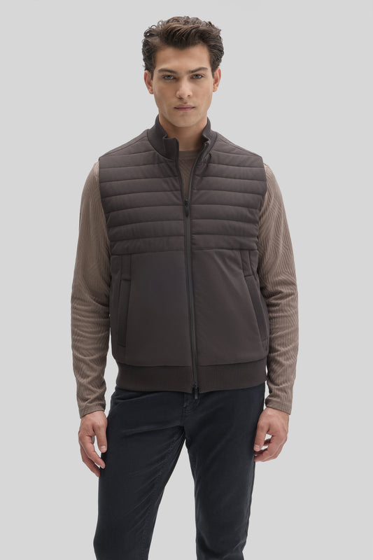 Man wearing dark gray quilted vest with horizontal stitching over beige long-sleeve shirt and dark pants.