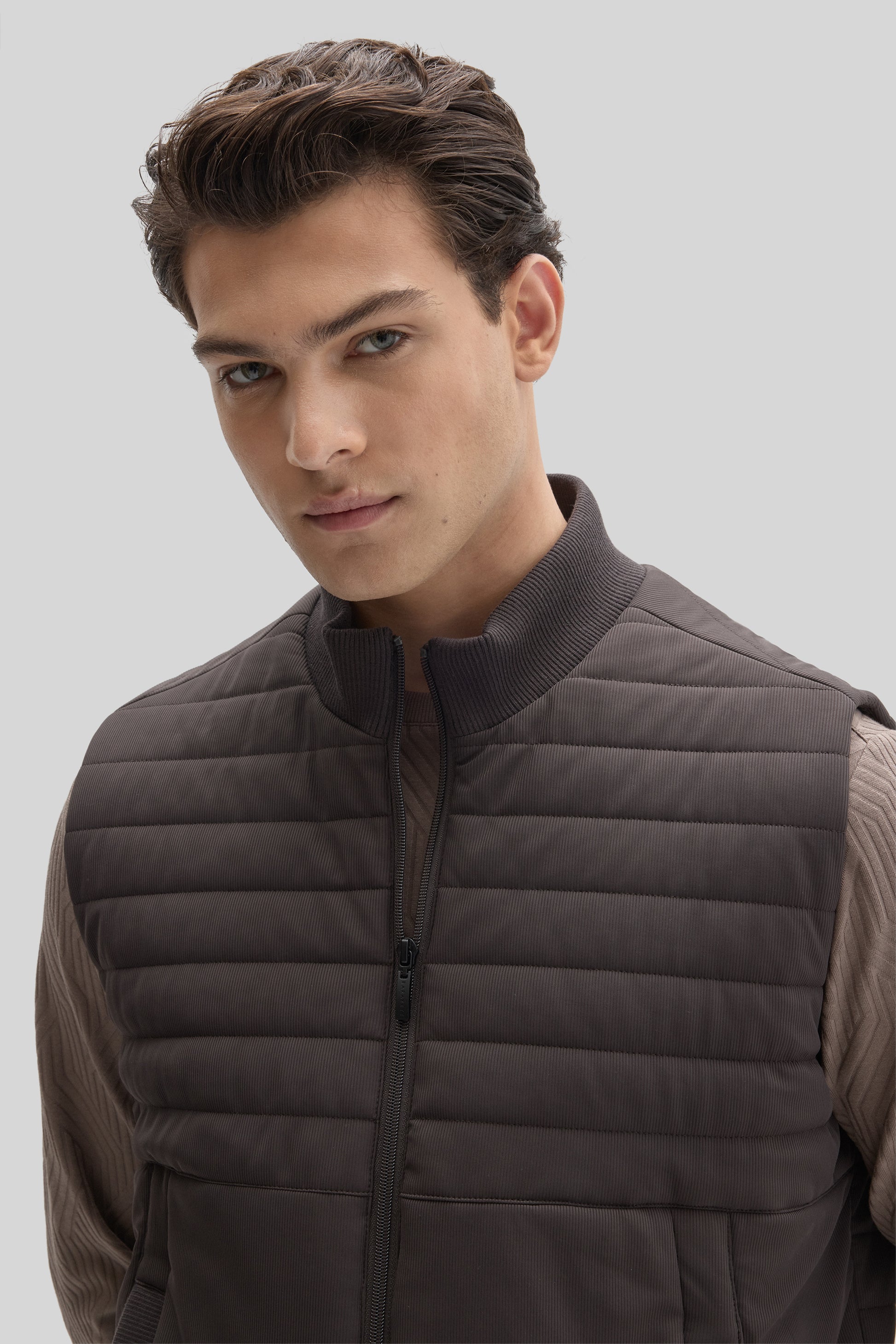 Man wearing a dark brown quilted vest with horizontal stitching over a ribbed long-sleeve shirt.