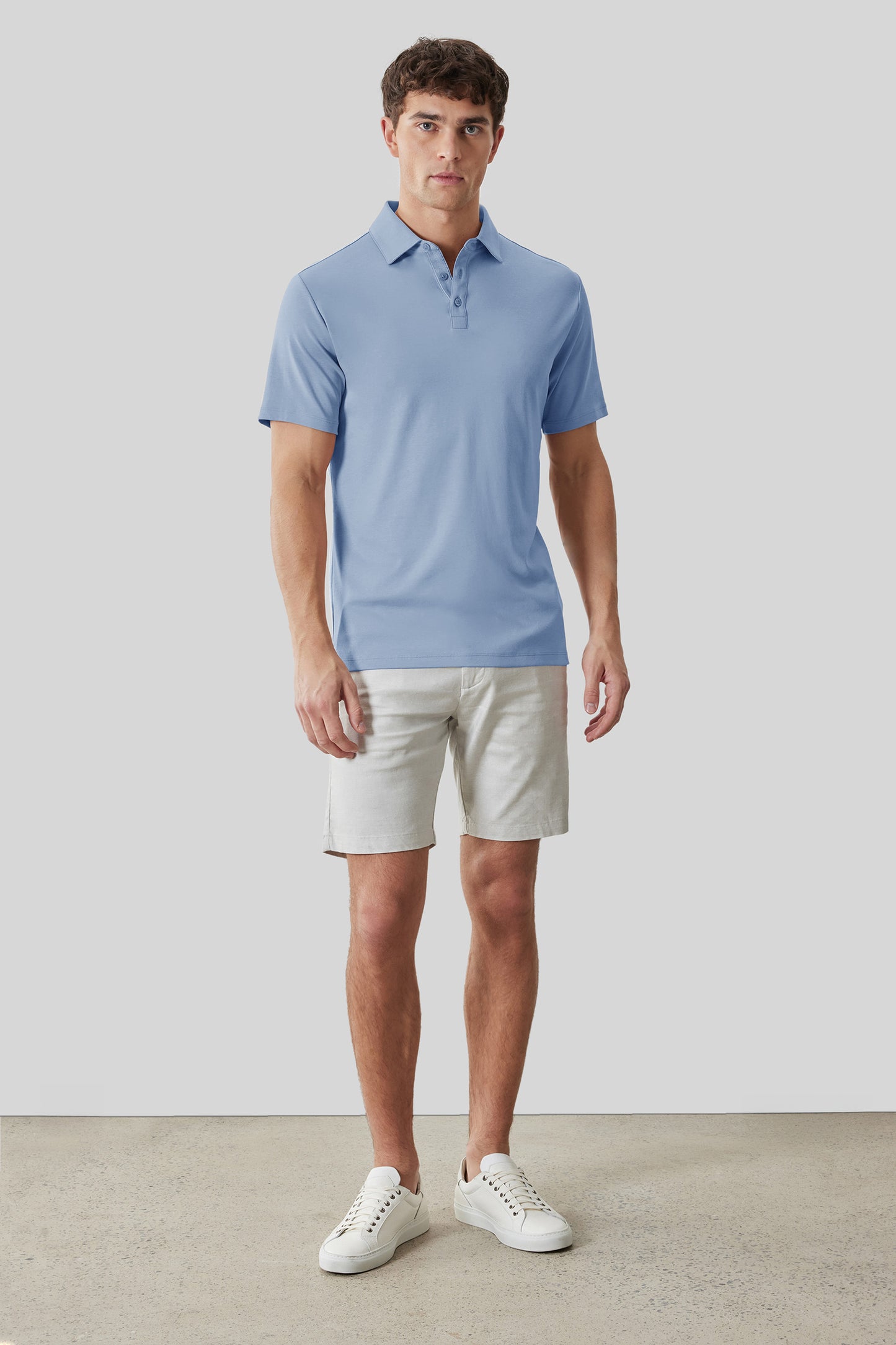 Man wearing light blue short-sleeve polo shirt with collar and buttons, paired with cream shorts and white sneakers.