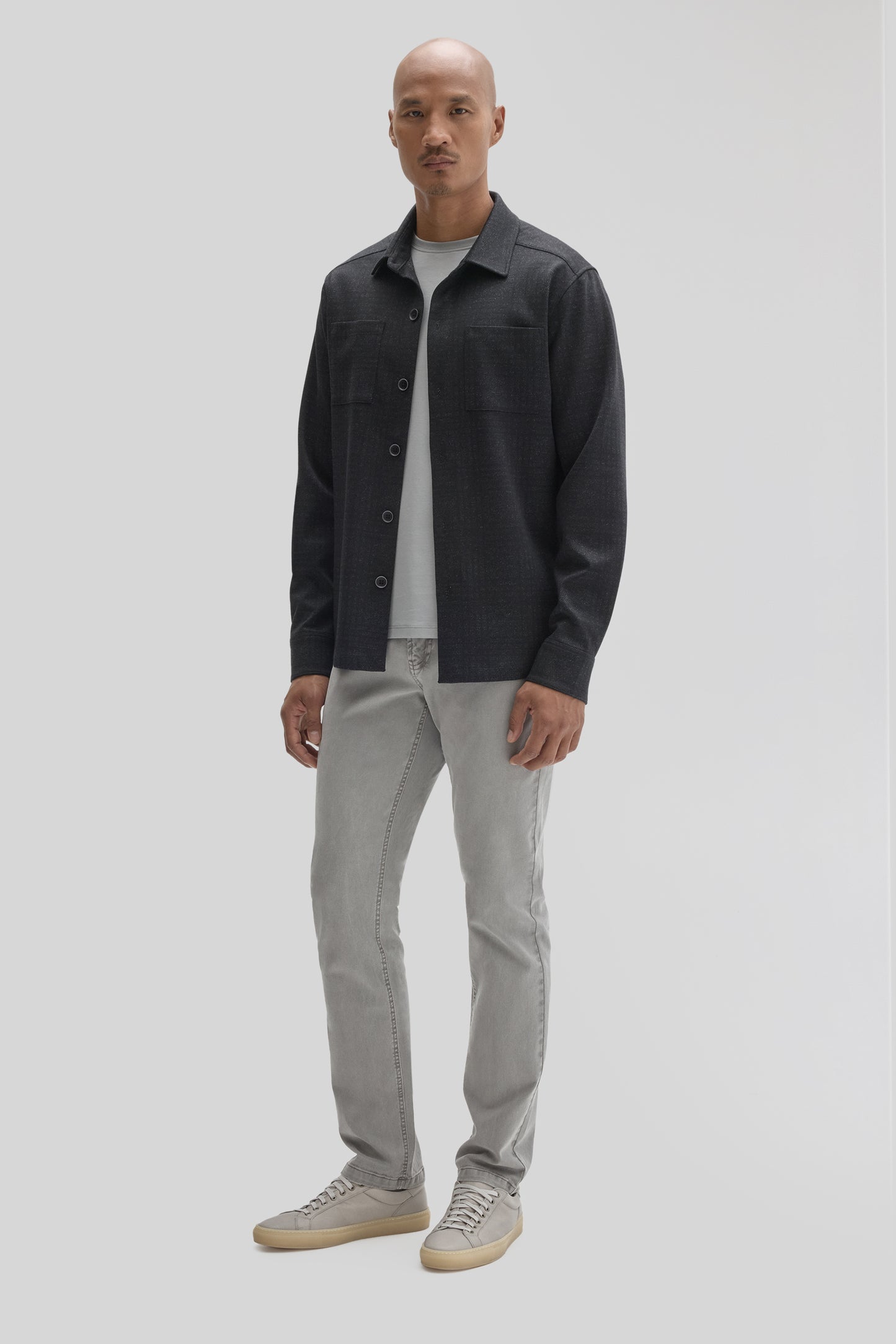 Man wearing charcoal button-up overshirt with chest pockets over white tee, light gray jeans, and gray canvas sneakers.