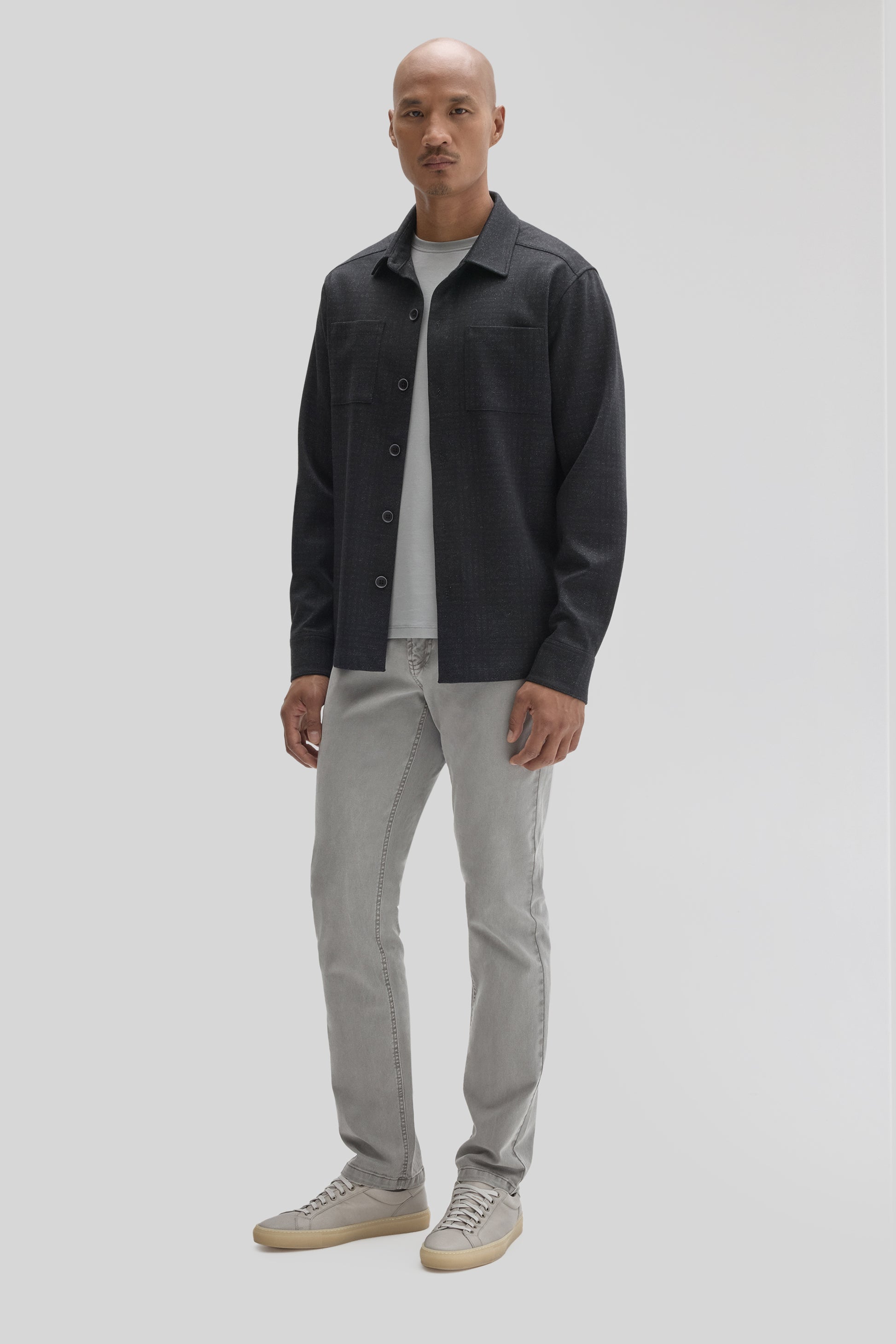 Man wearing charcoal button-up overshirt with chest pockets over white tee, light gray jeans, and gray canvas sneakers.
