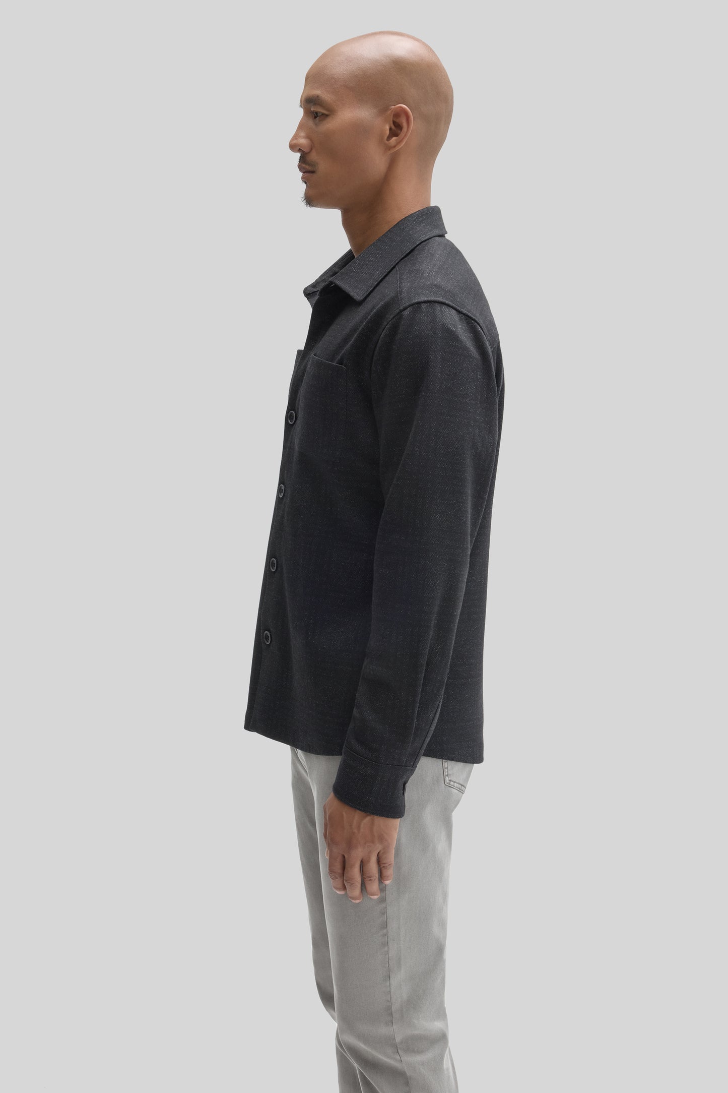 Man wearing dark charcoal overshirt with collar and button front over light gray pants, shown in profile.