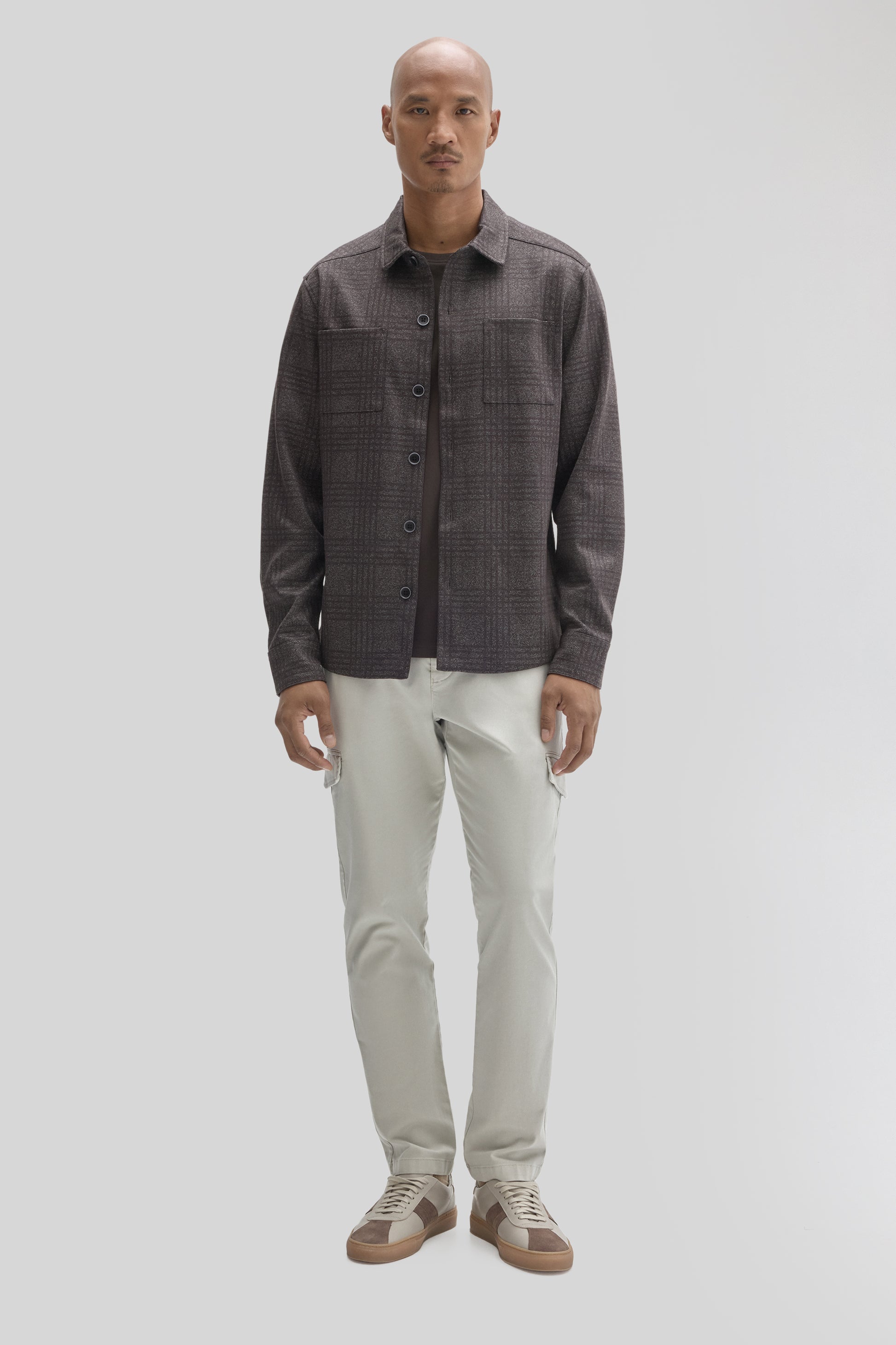 Man wearing charcoal textured overshirt with chest pockets and button front over cream pants and tan sneakers.