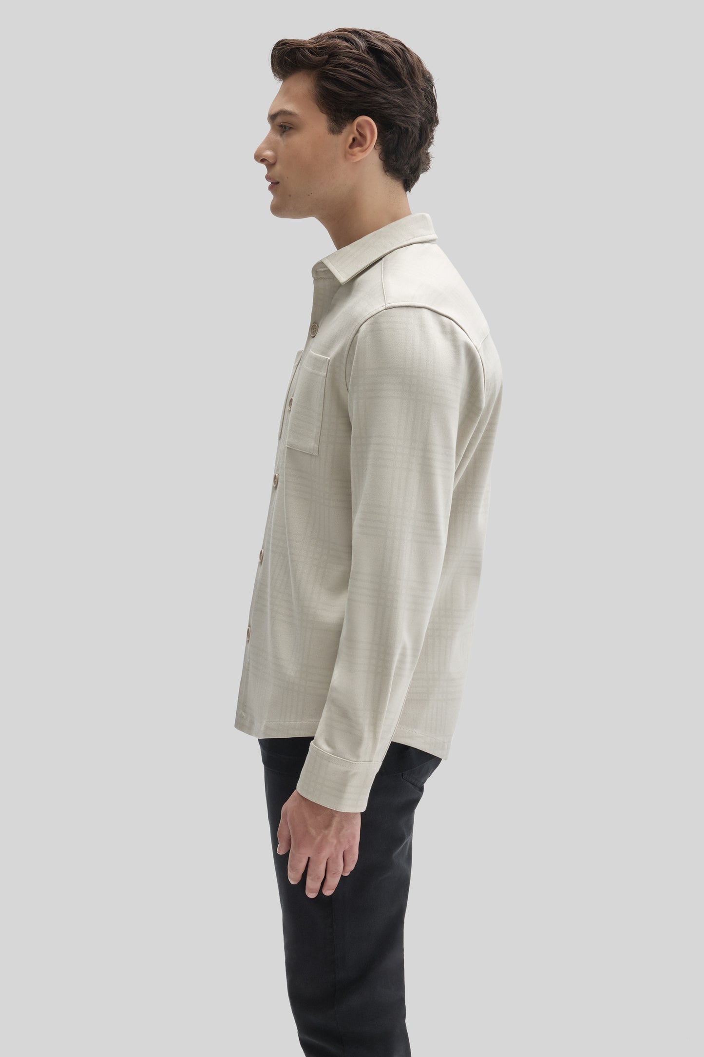 Man wearing cream-colored button-up overshirt with collar and chest pocket over black pants, shown in side profile.