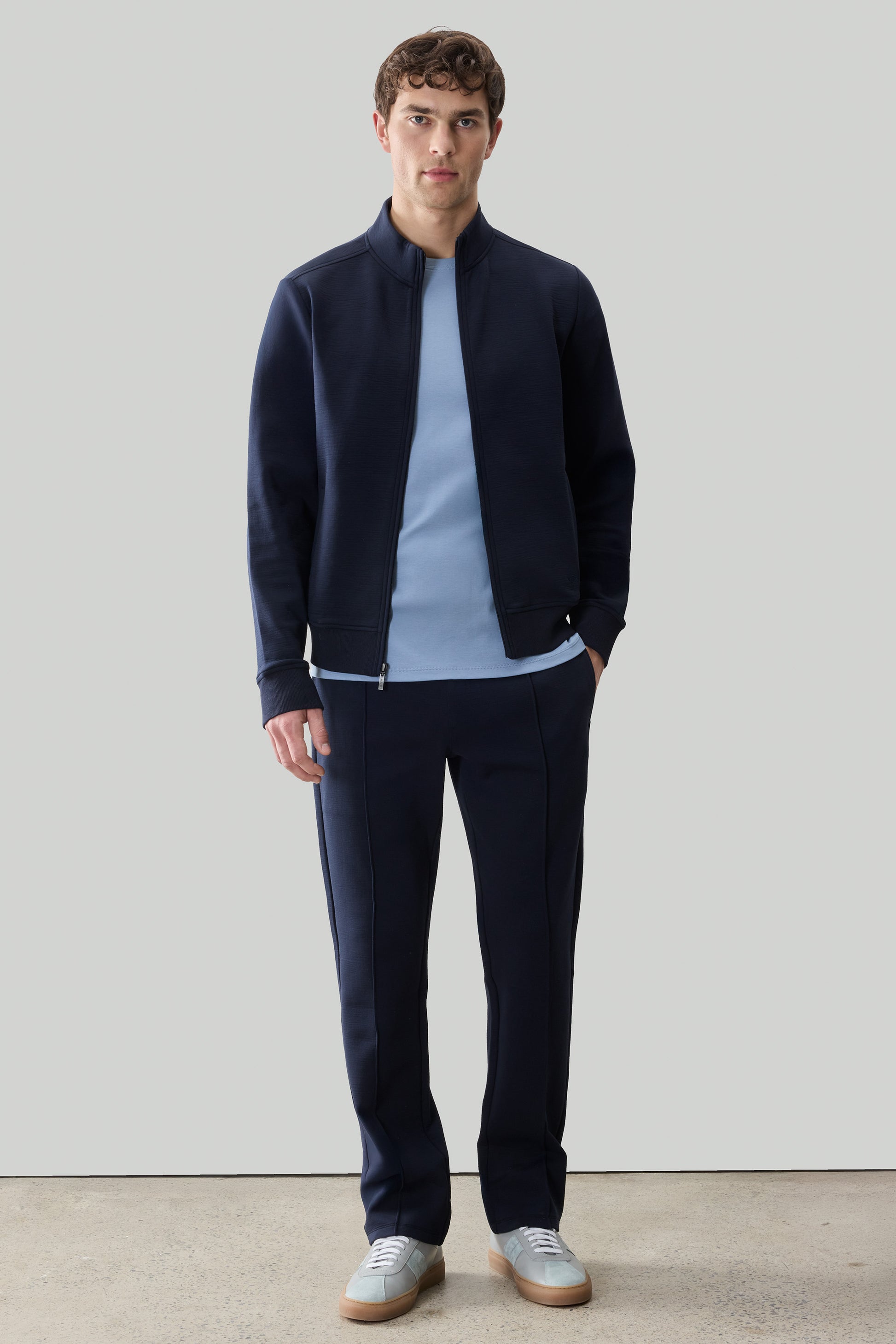 Man wearing navy full-zip long sleeve jacket over light blue t-shirt and navy pants with white sneakers.