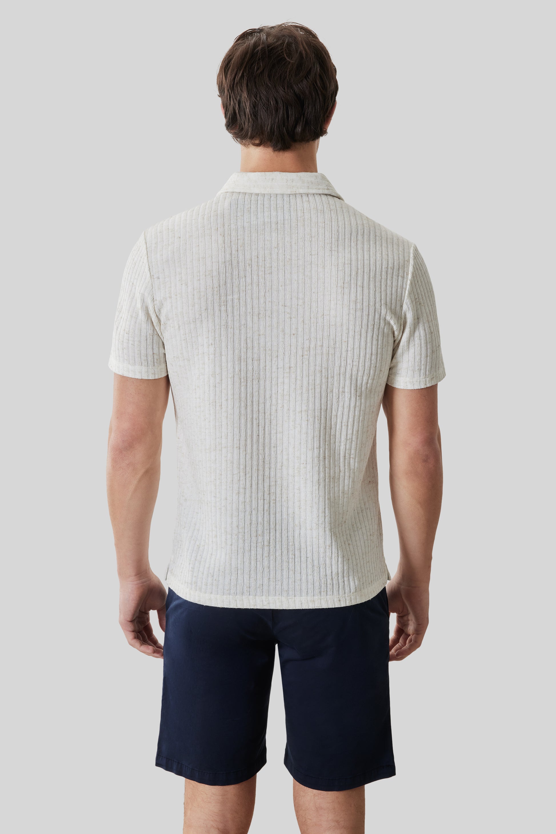 Man wearing cream ribbed short-sleeve polo shirt with open collar and dark navy shorts, shown from back.