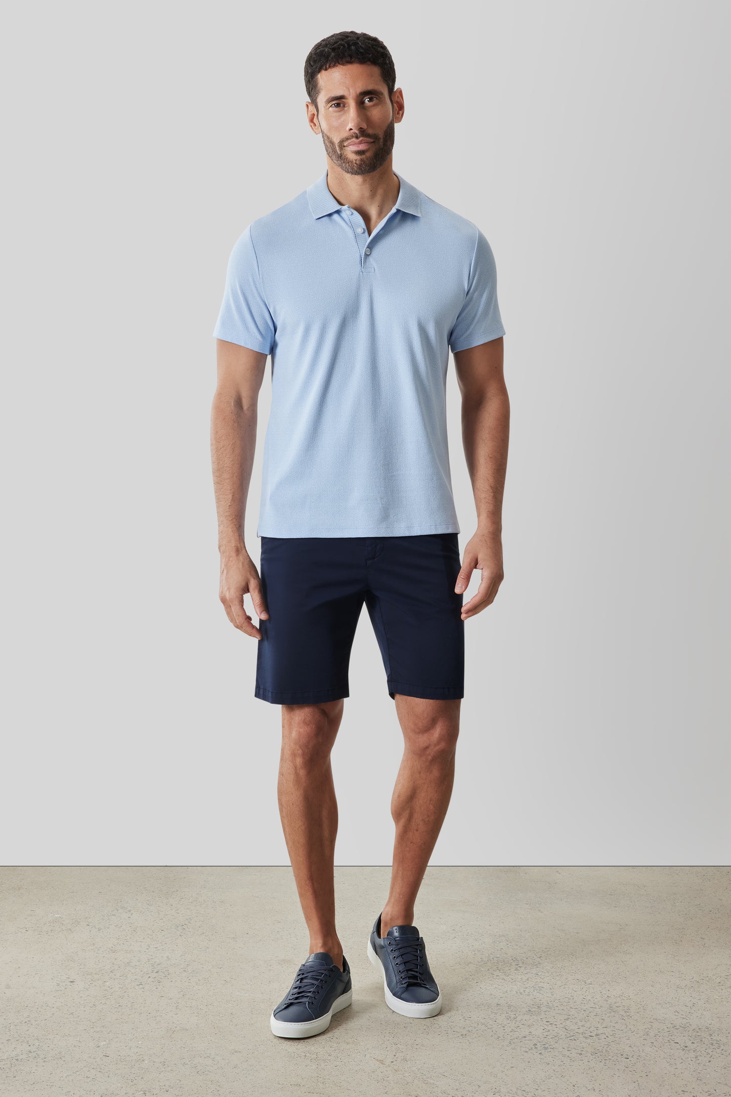 Man wearing light blue short-sleeve polo shirt with dark navy shorts and navy sneakers.