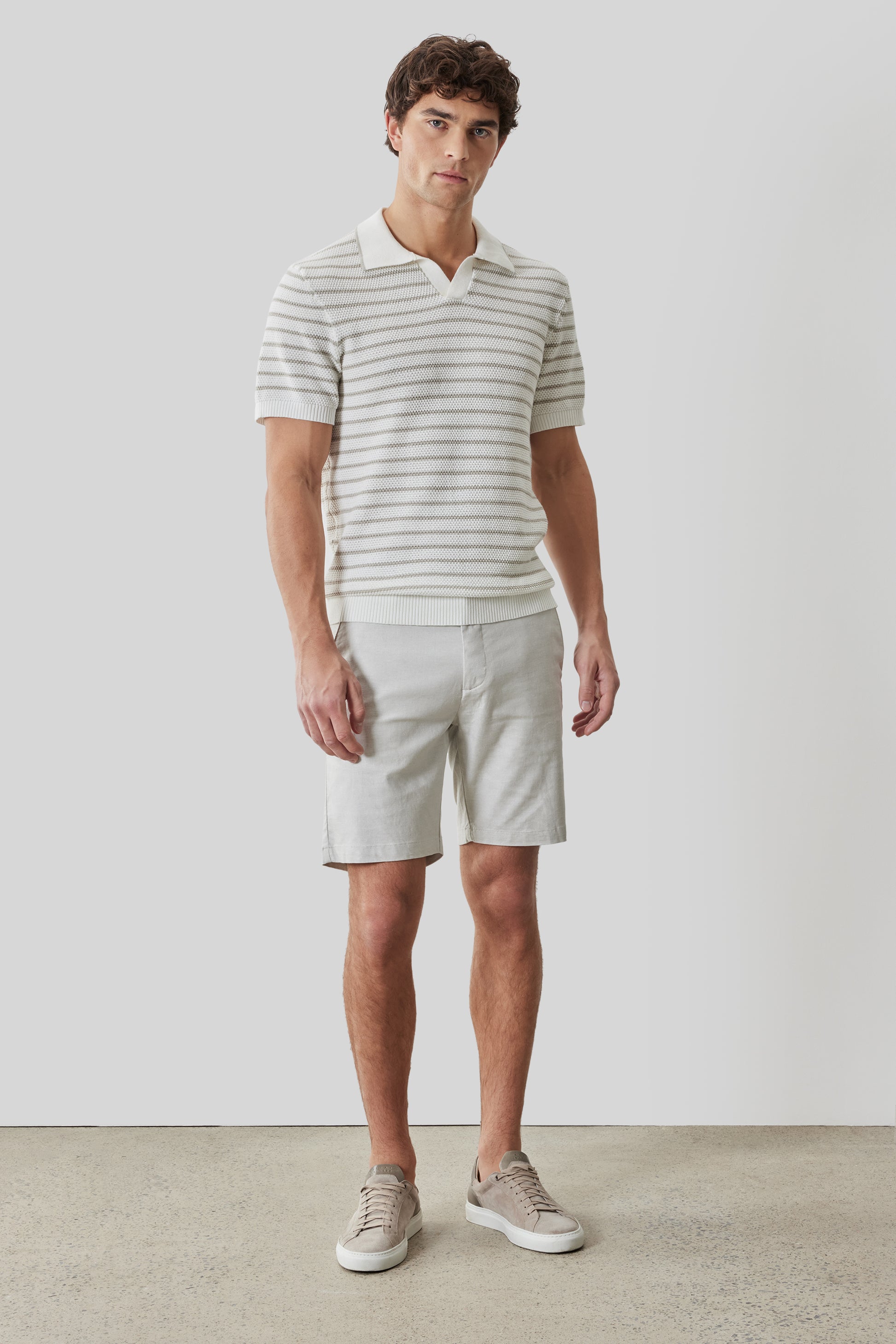 Man wearing striped short-sleeve polo shirt with open collar in cream and gray, paired with beige shorts and sneakers.