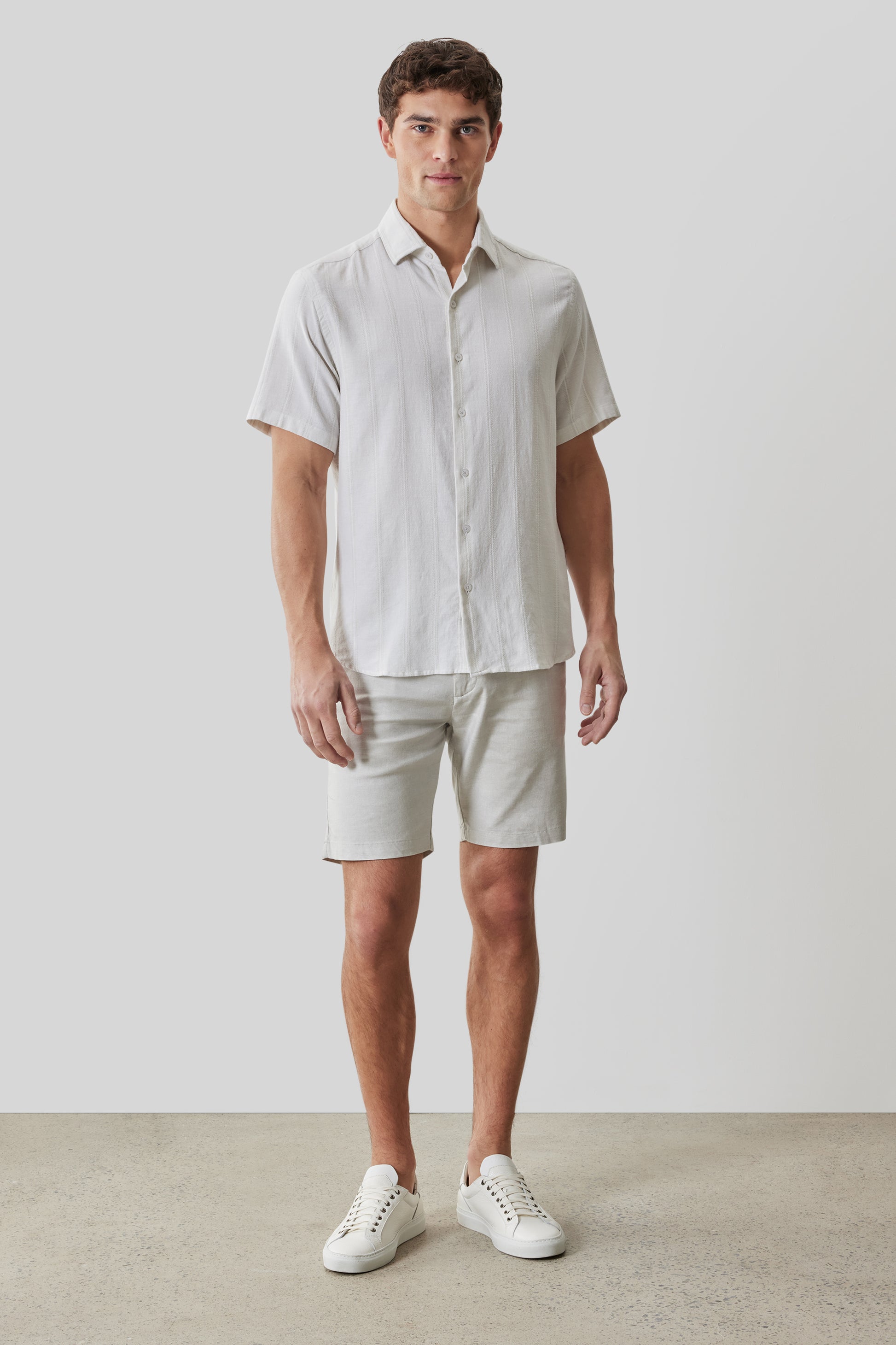 White short-sleeve button-up shirt with vertical texture, worn with cream shorts and white sneakers.
