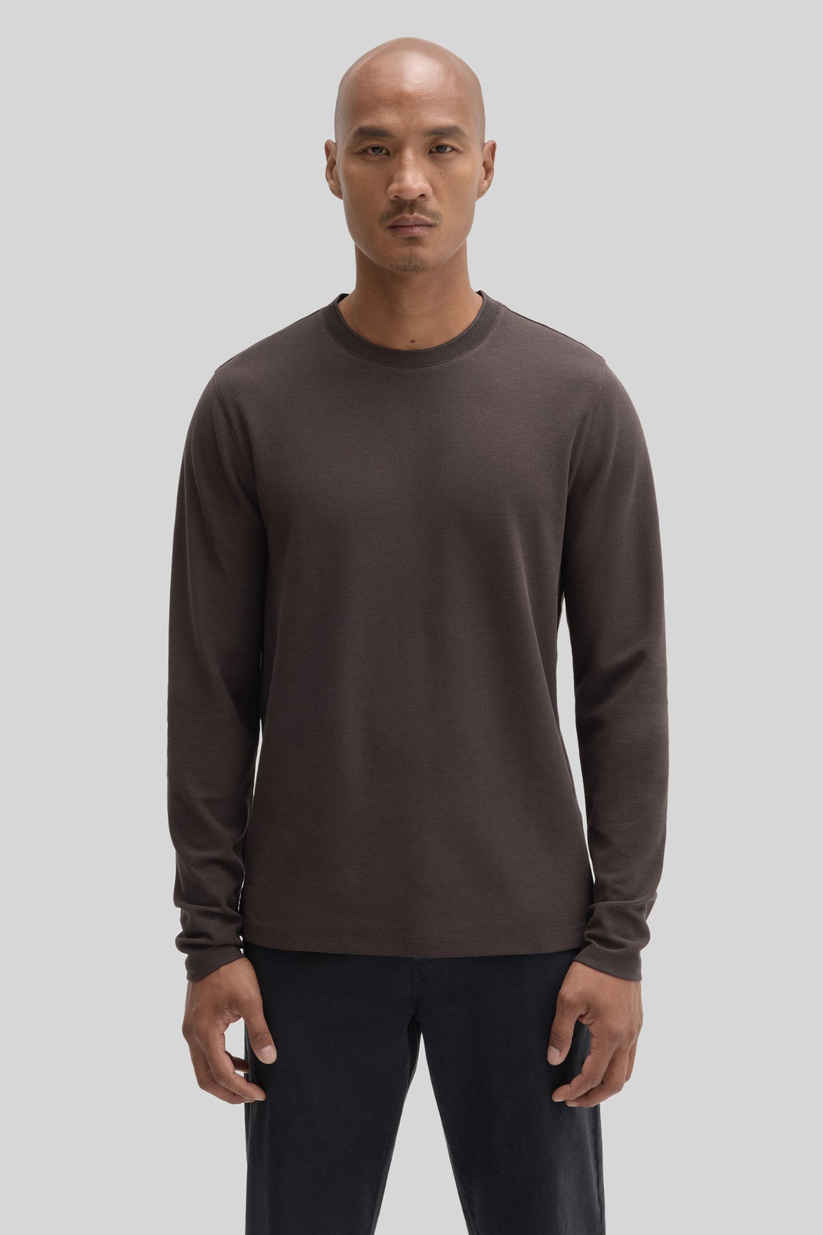 Man wearing a charcoal long sleeve crew neck shirt with dark pants against a neutral background.