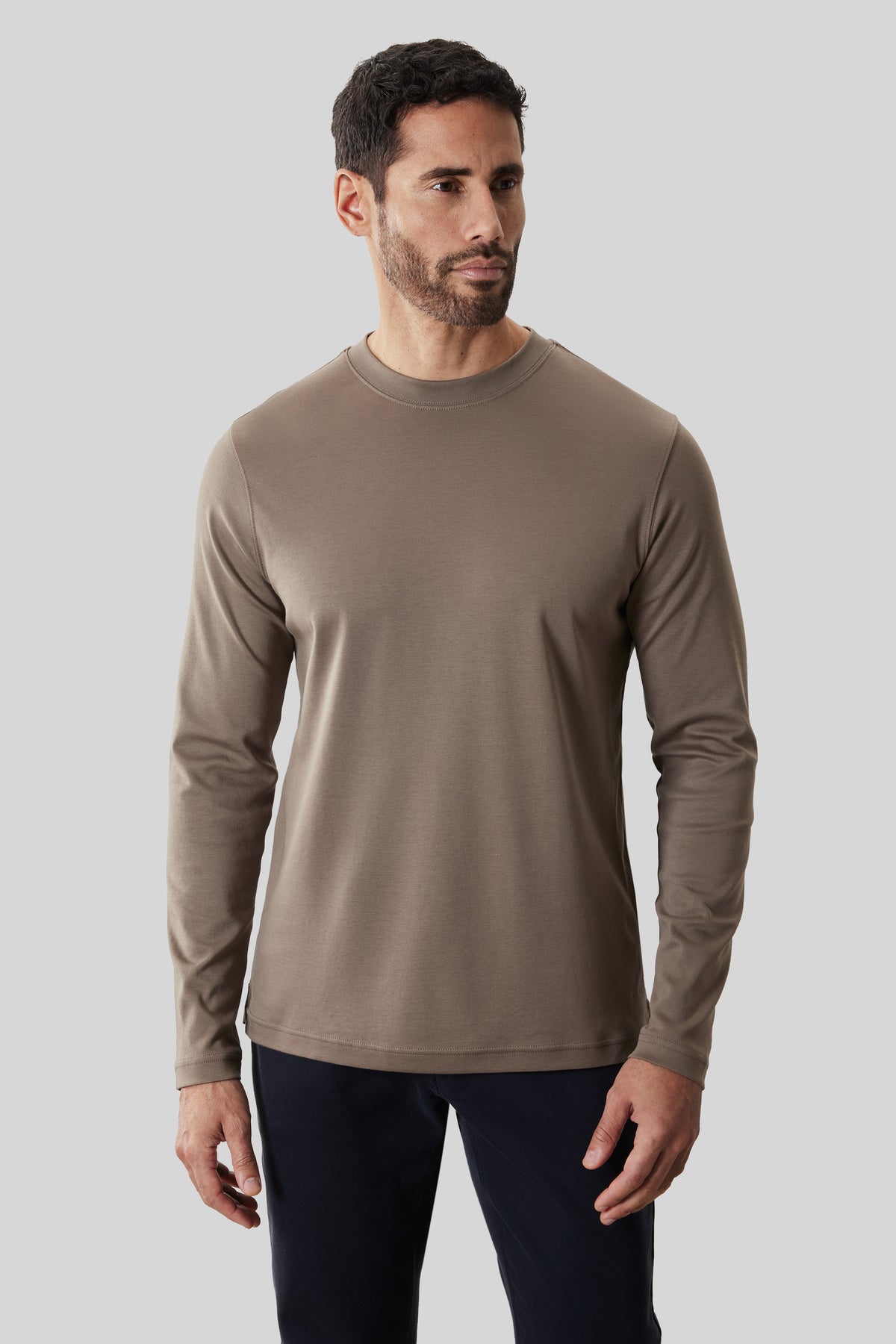 Man wearing taupe long-sleeve crew neck shirt with dark navy pants against neutral background.