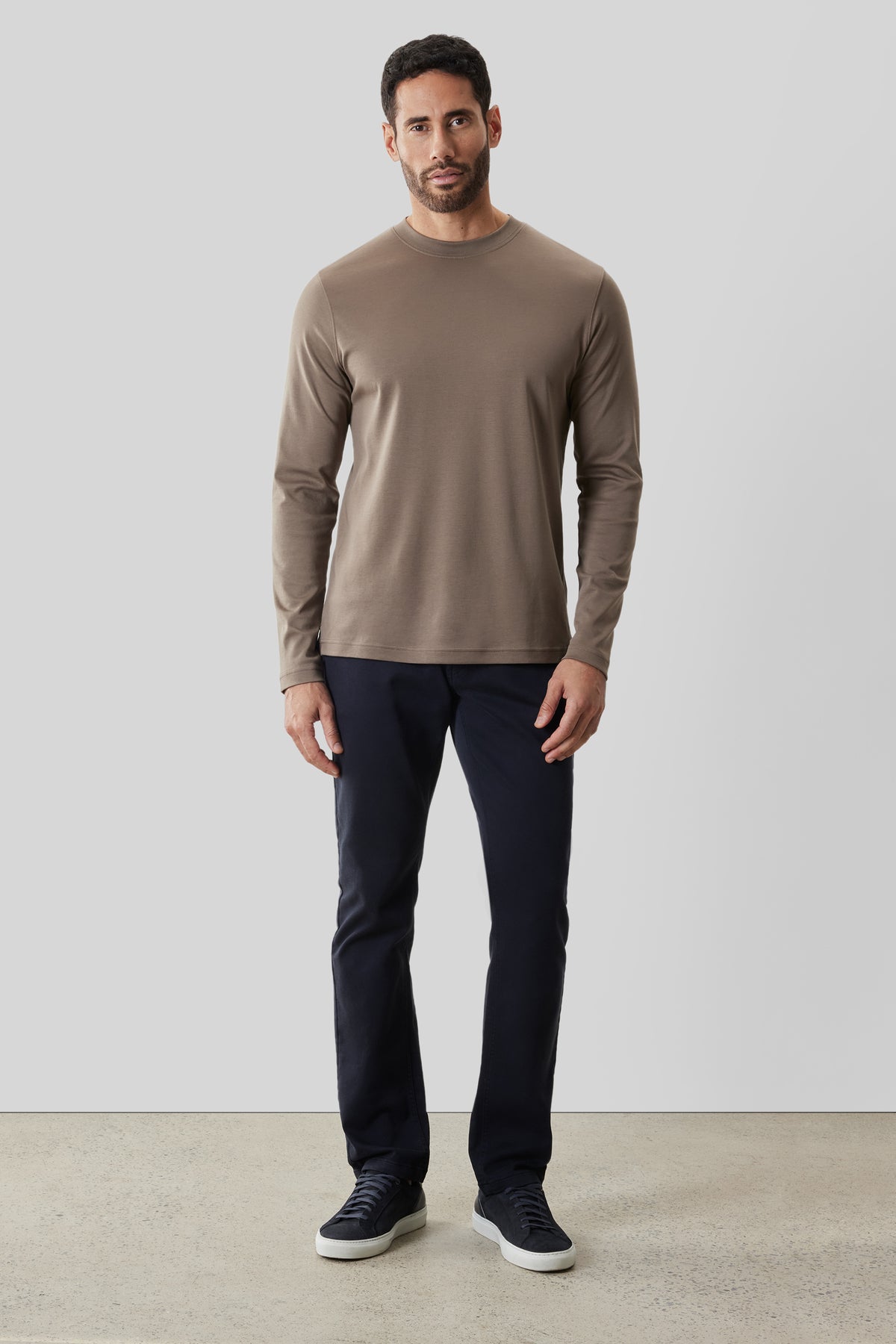 Man wearing taupe long-sleeve crew neck shirt with dark navy pants and dark sneakers.