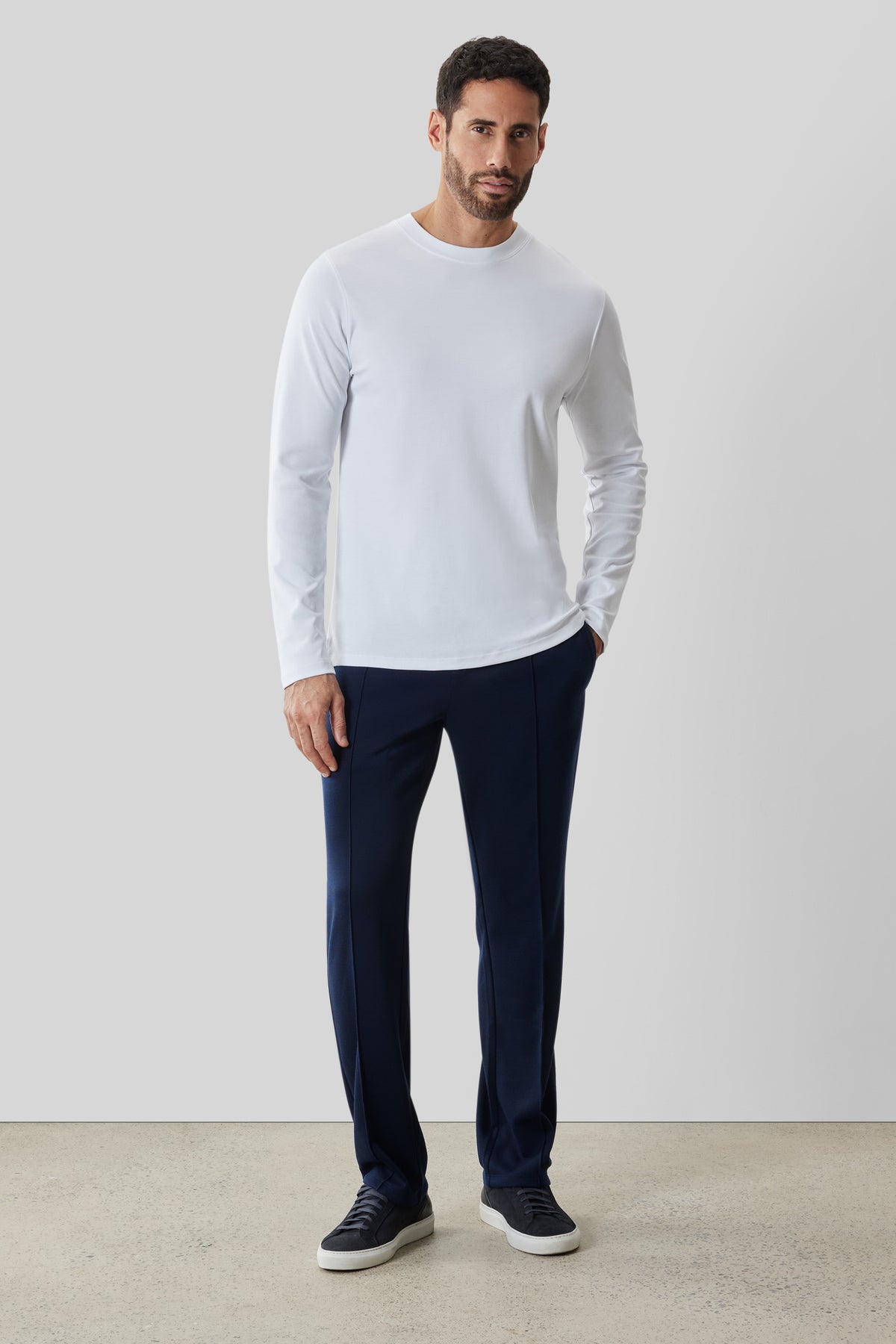 Man wearing white long-sleeve crew neck shirt with navy pants and black sneakers.