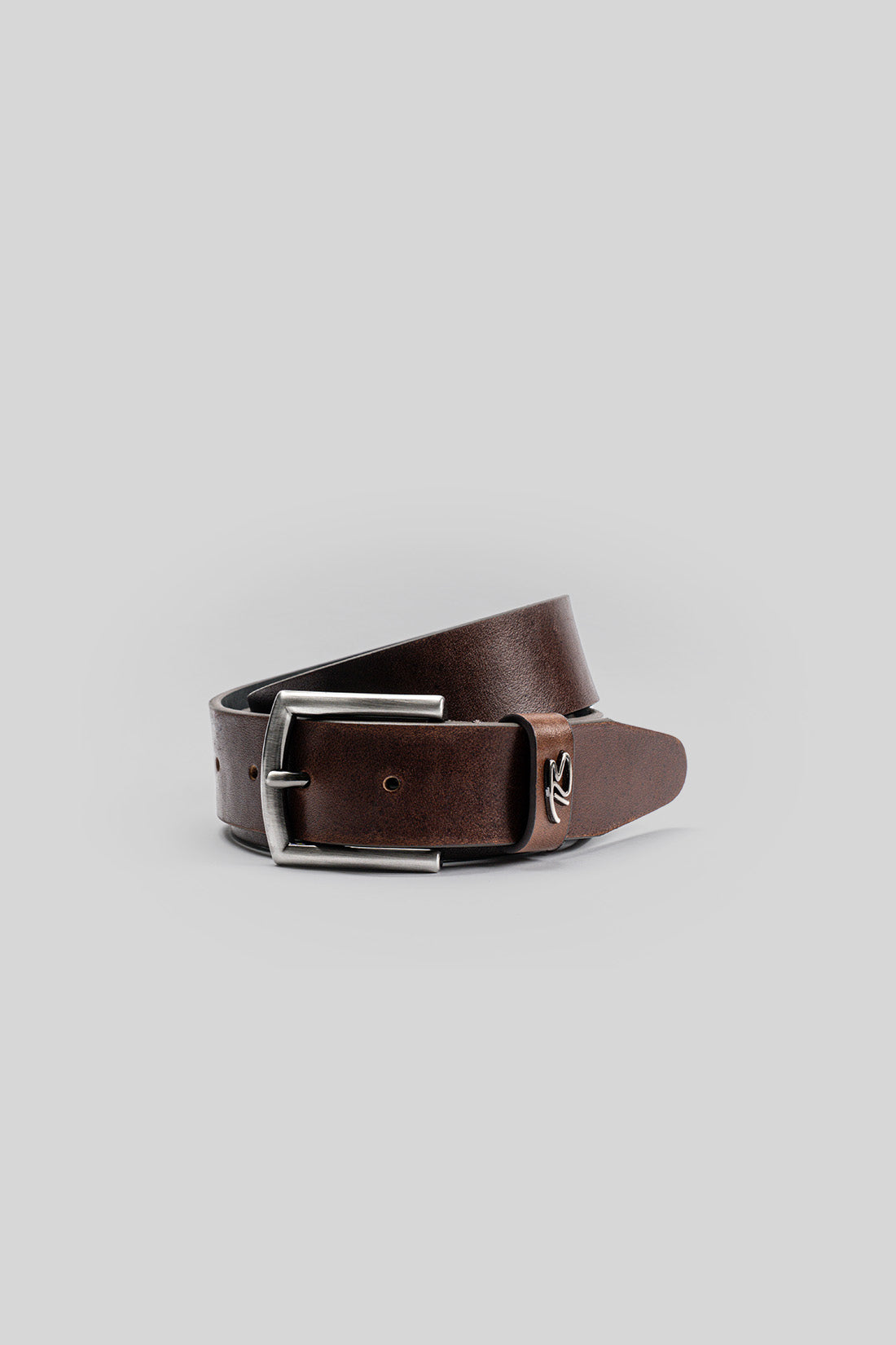 Brown leather belt with silver rectangular buckle and metal side emblem on gray background.