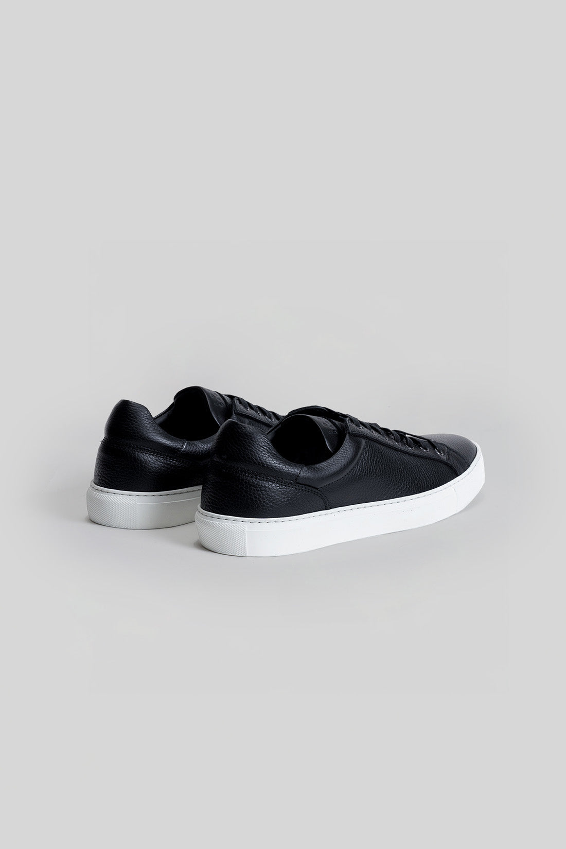 Black leather sneaker with white rubber sole, shown from rear three-quarter view.