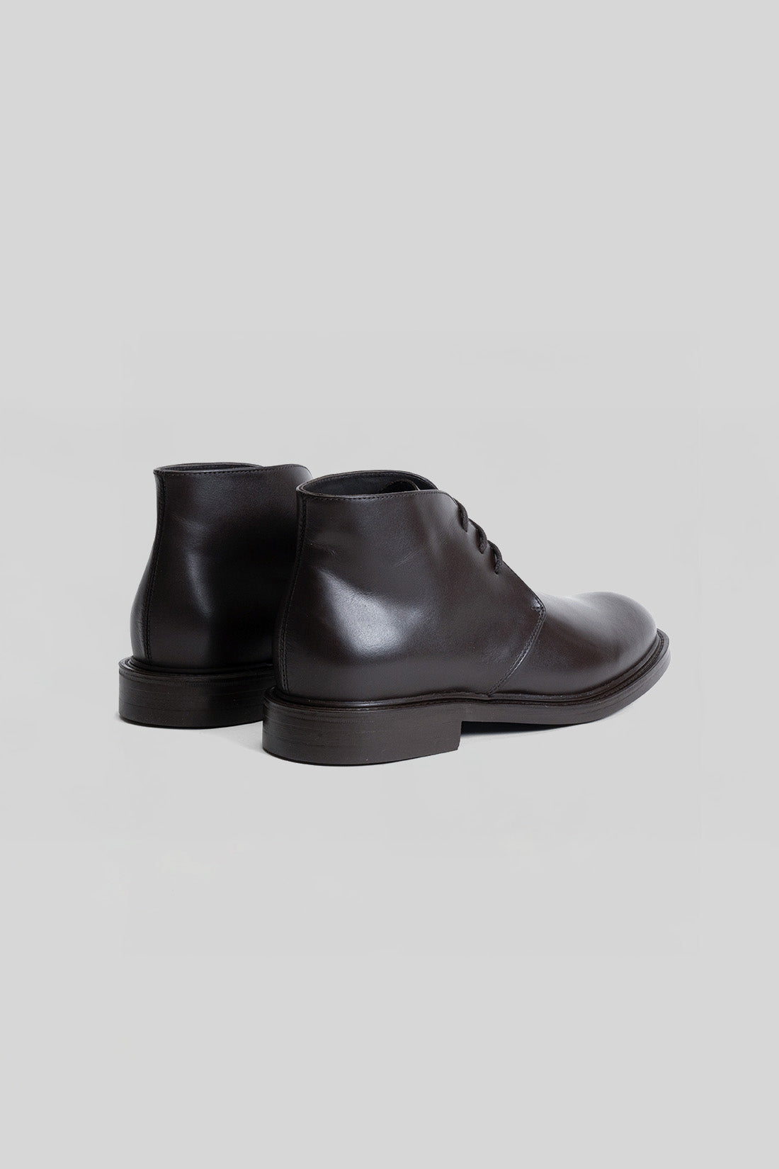 Black leather chukka boot with lace-up front and flat sole, shown from rear three-quarter view.
