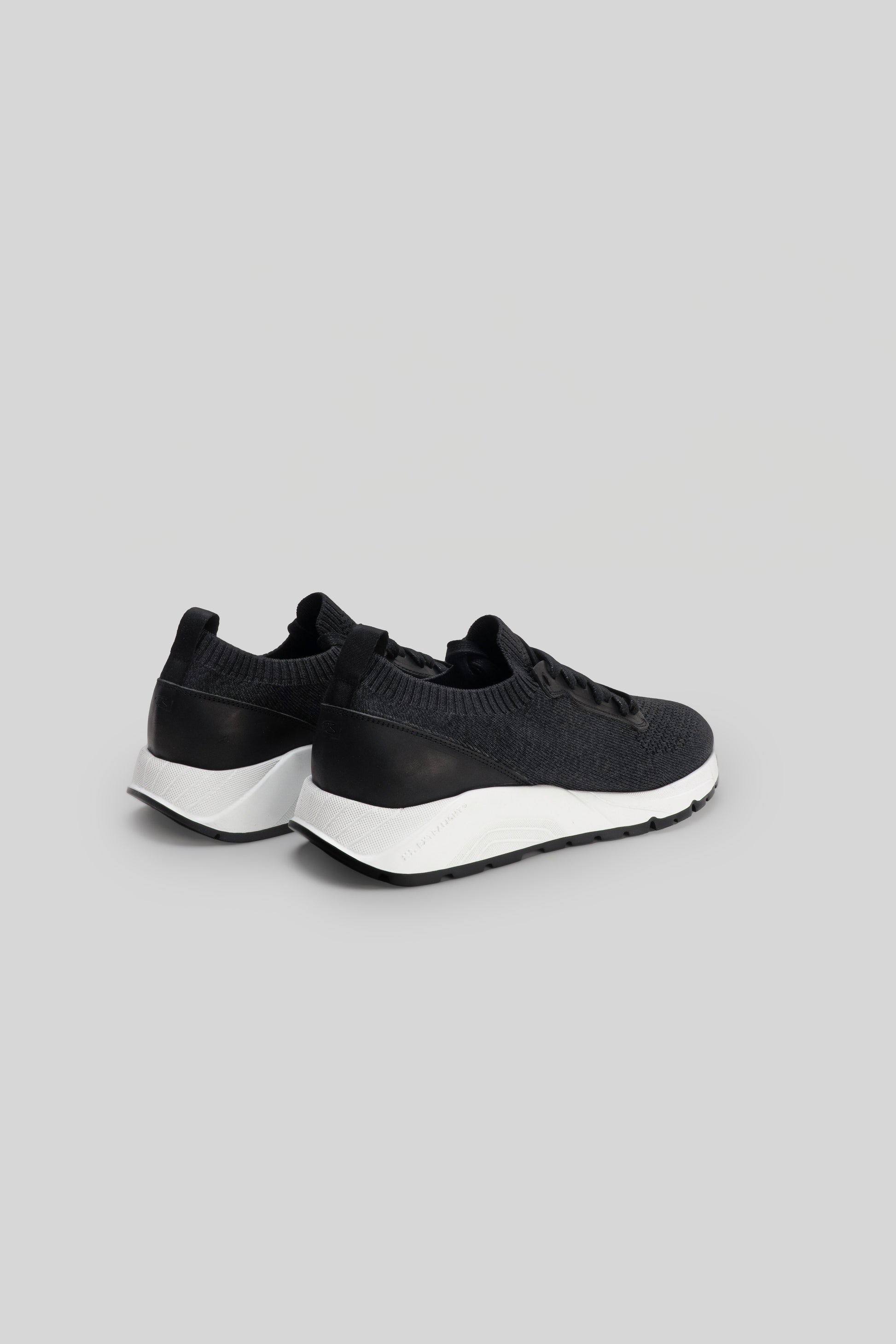 Black knit sneaker with white sole and black heel panel, shown from rear three-quarter view.