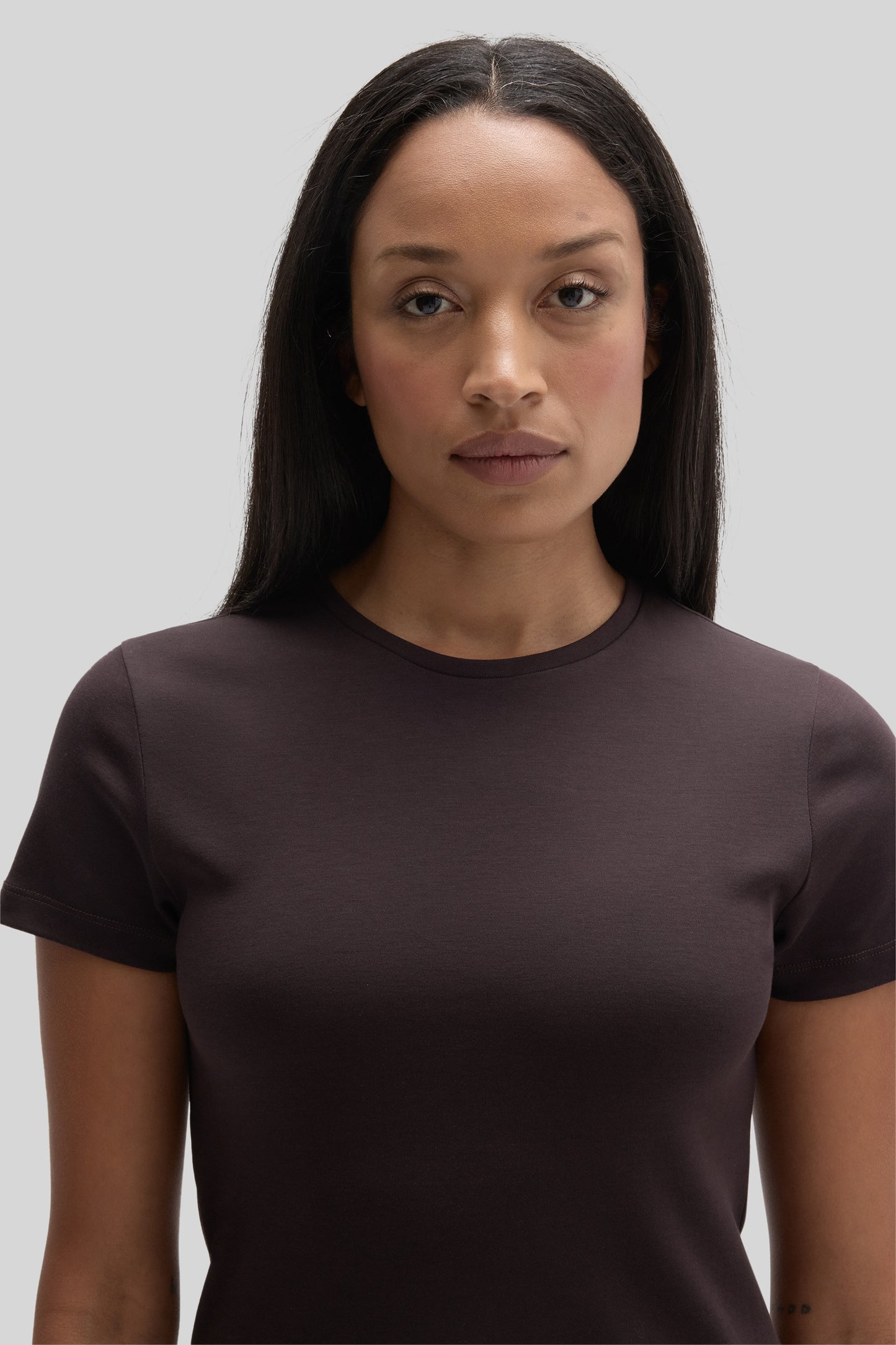 Woman wearing a dark brown crewneck short-sleeve t-shirt against a gray background.